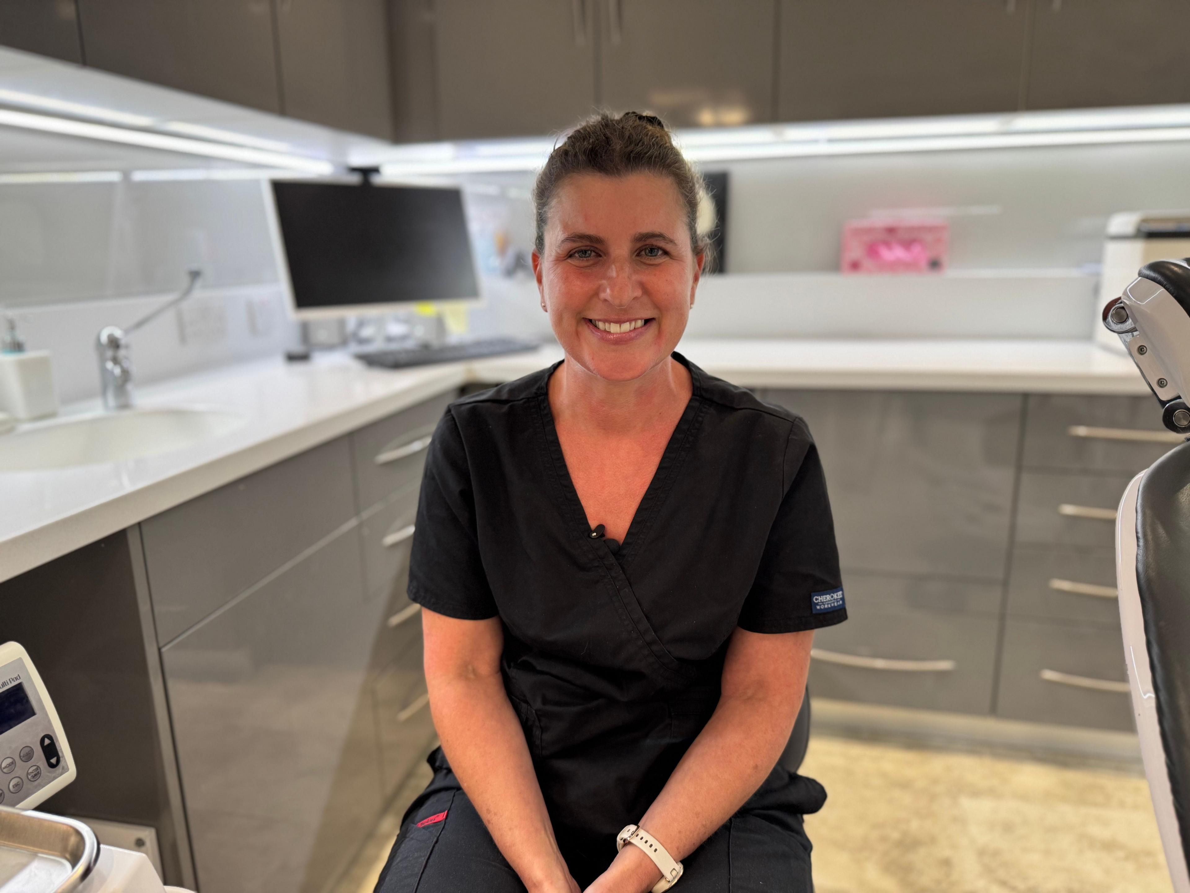 A woman in her 30's in black dentist scrubs. She wears her hair tightly pulled back and she is smiling at the camera.