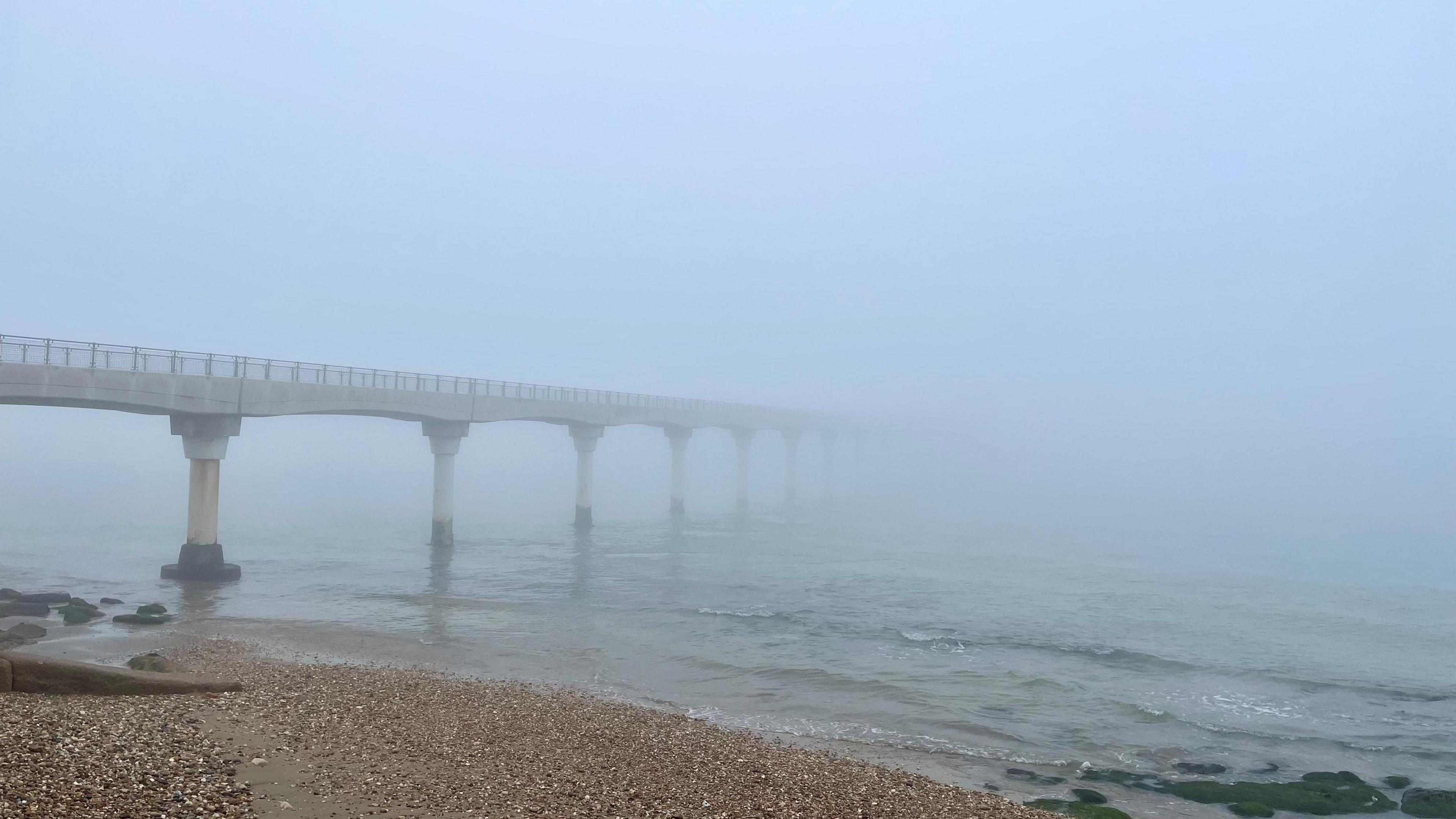 A very foggy coastal scene.  A pier vanishes into the fog at Bembridge. Visibility is very limited and the sky is white. In the foreground you can see a pebble beach and waves gently lapping at the shore.