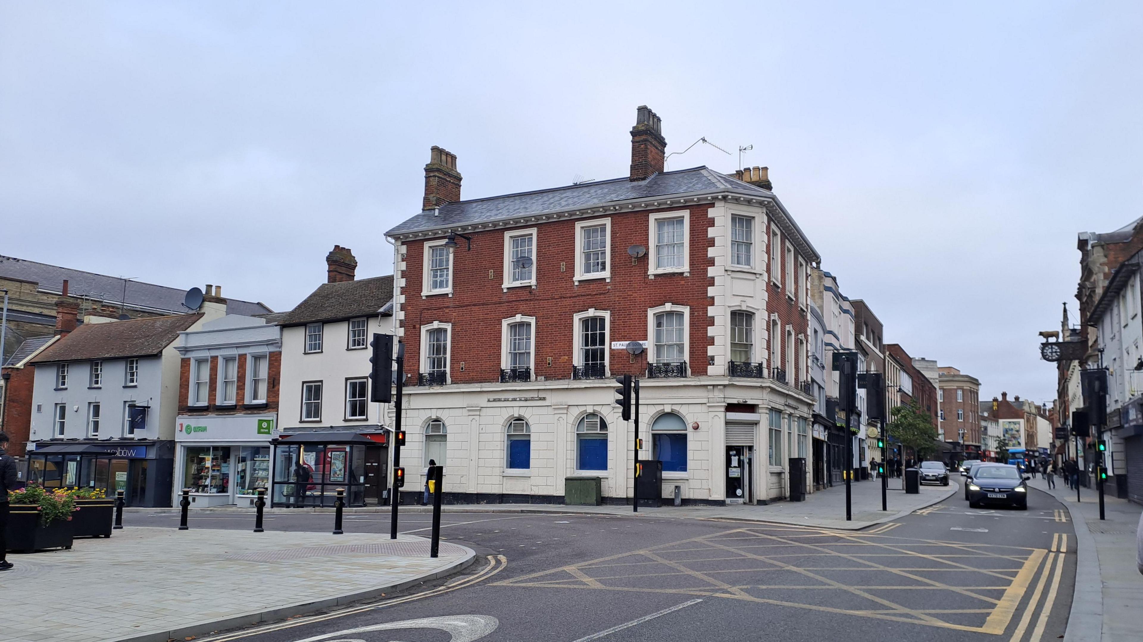 An scene featuring a red-brick corner building with white accents, surrounded by shops and businesses. Road markings and pedestrian signals are visible while people stroll along the pavements. 