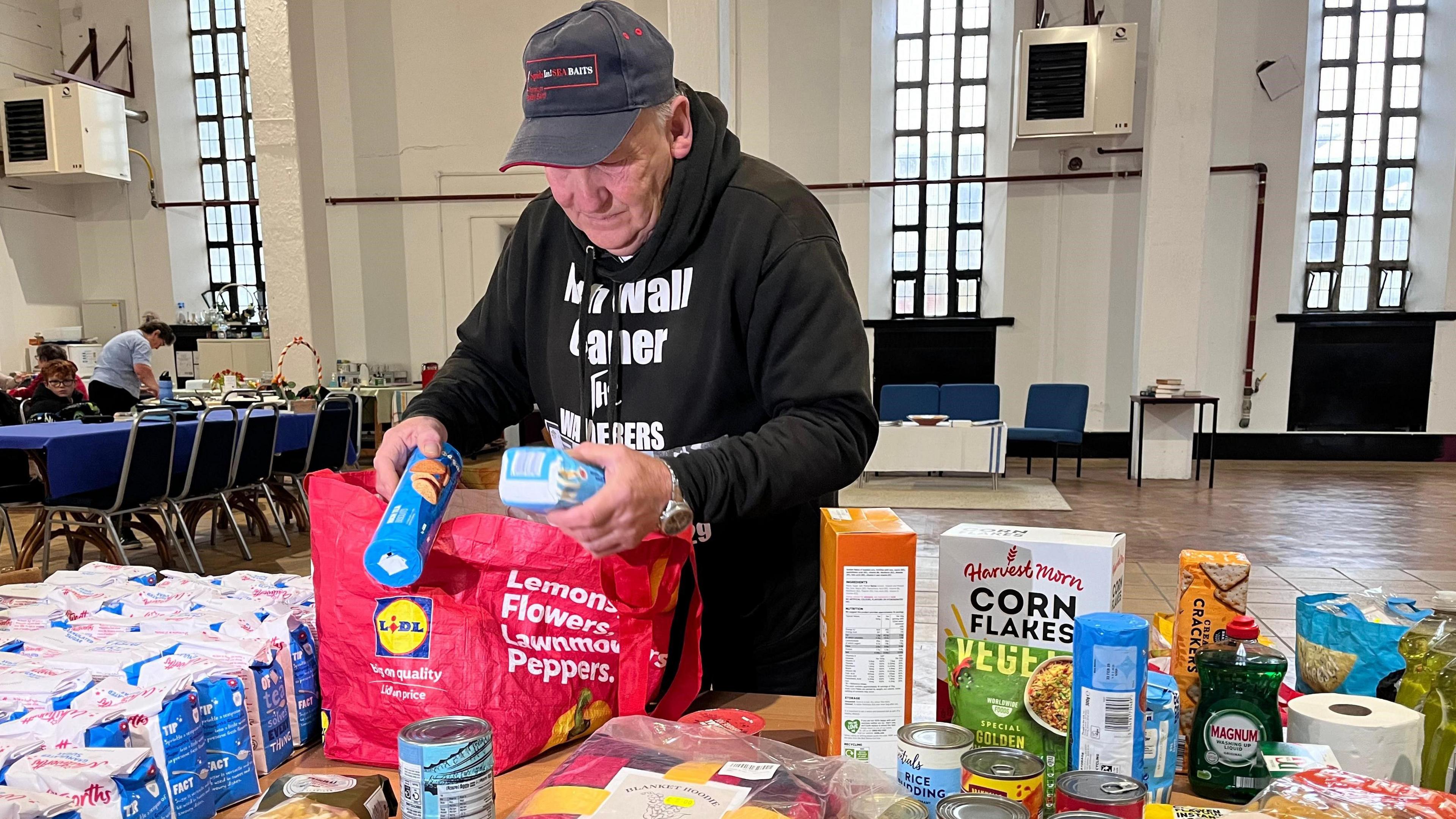 Frank Sparkes wearing a black hoodie and cap packing food items, including sugar, cornflakes and canned goods, into a bag