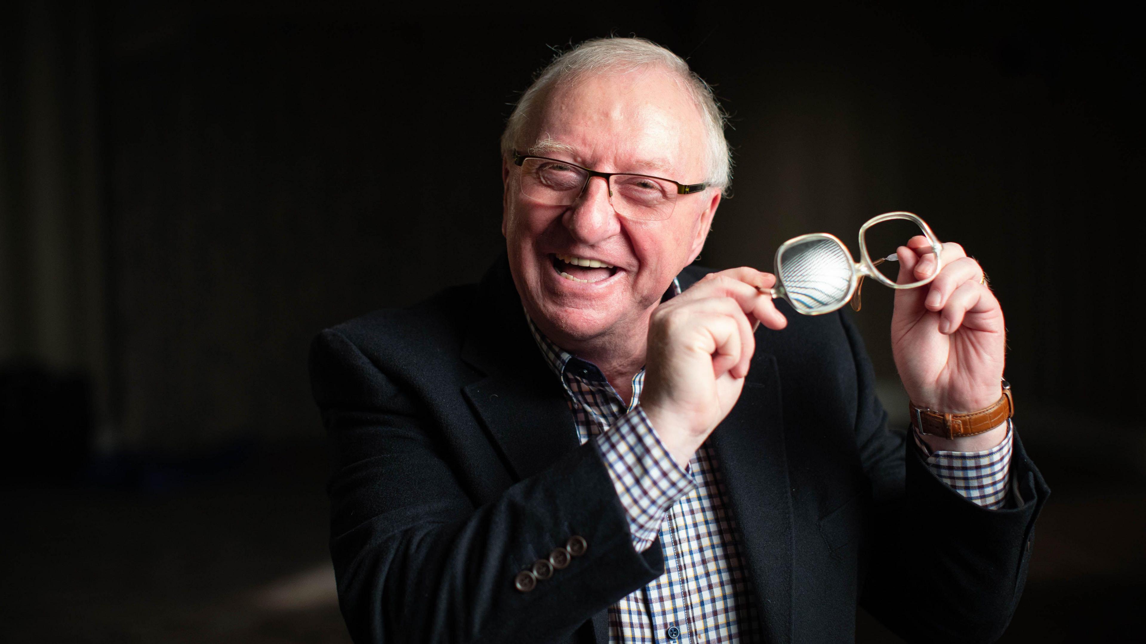 Dennis Taylor, holding the pair of glasses he wore during the famous snooker match in 1985. He is wearing a new set of glasses and a black jacket of a check shirt.