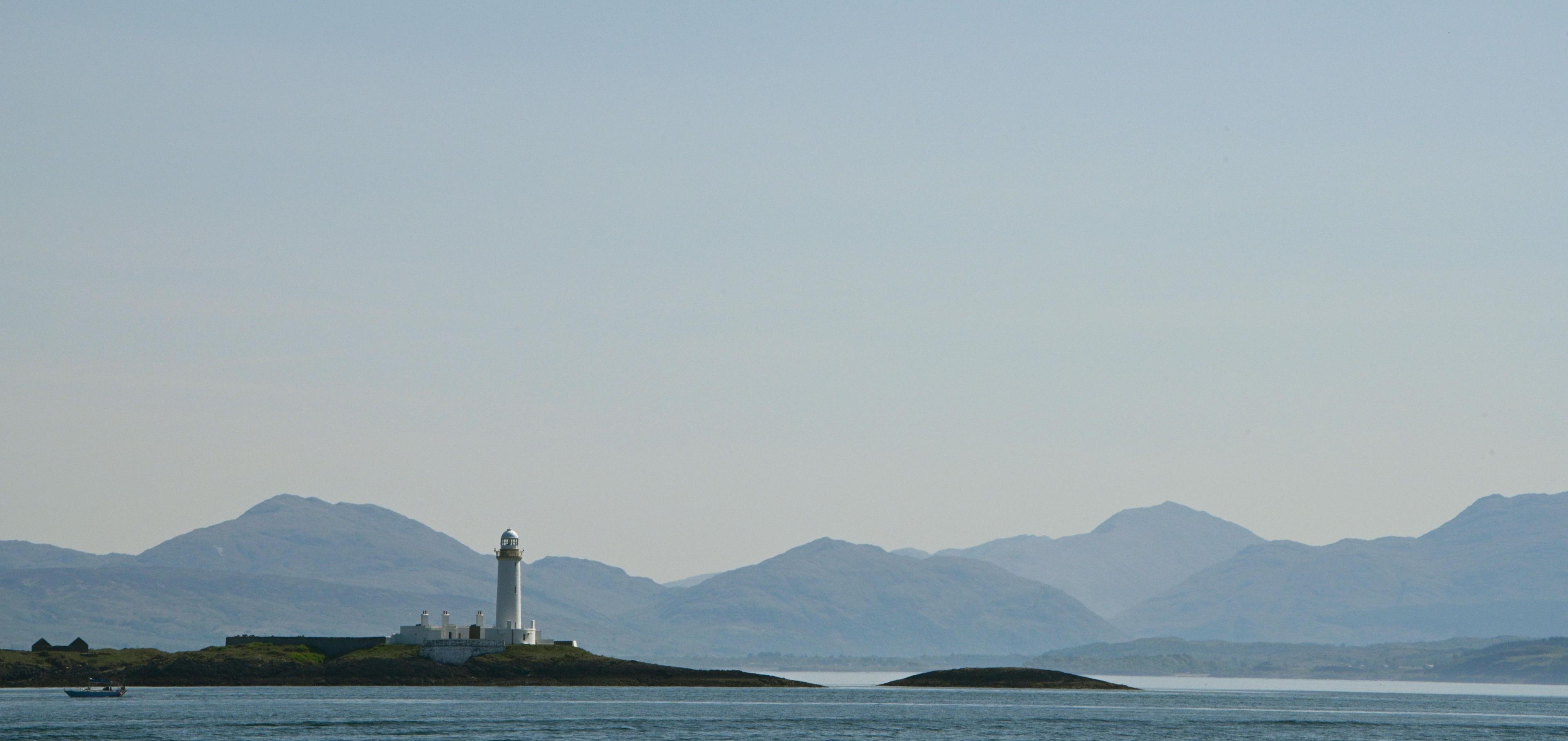 Landscape image with blue and grey tones of a lighthouse and calm waters with hills partially covered by fog in the far distance