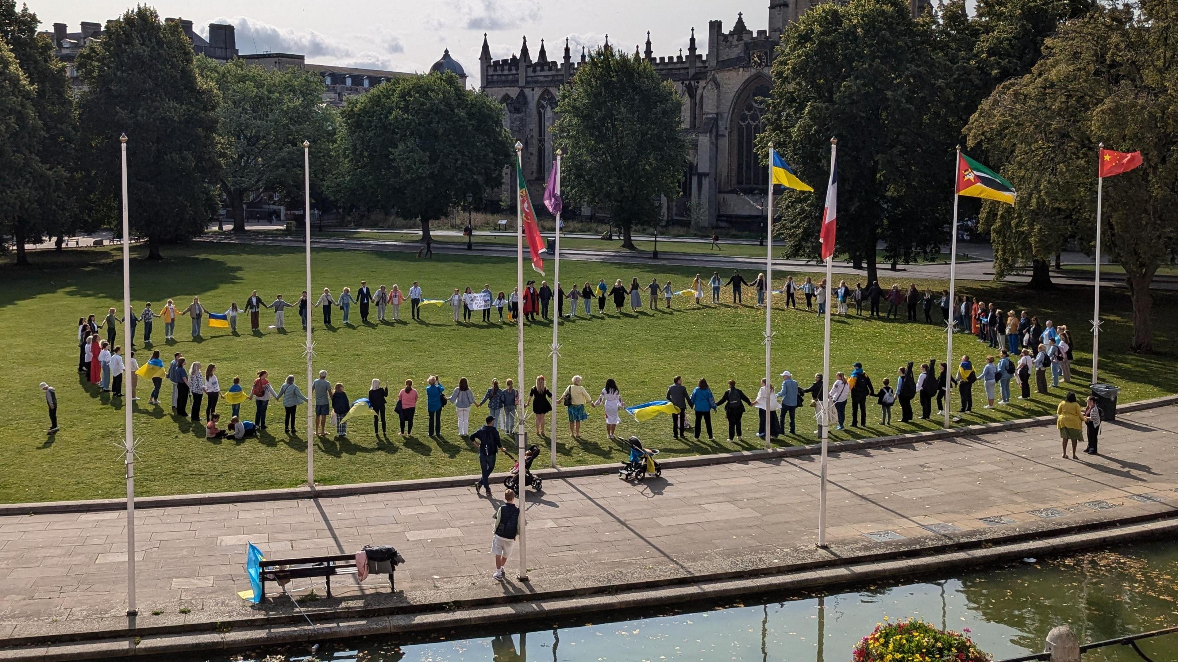 A ring of people holding hands standing on grass on College Green, with the photograph taken from a slightly raised position. Multiple flags are visible including the Ukrainian flag