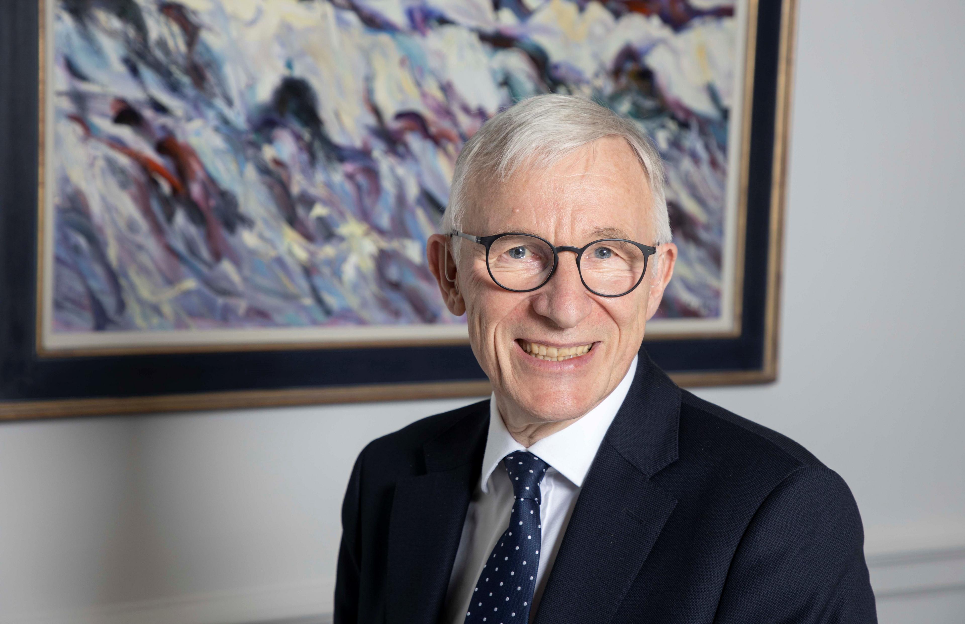 Lord Brodie smiles at the camera while standing in front of an abstract painting. He has white hair and is wearing a dark blue blazer, a white shirt, a dark blue spotted tie and round-rimmed glasses.