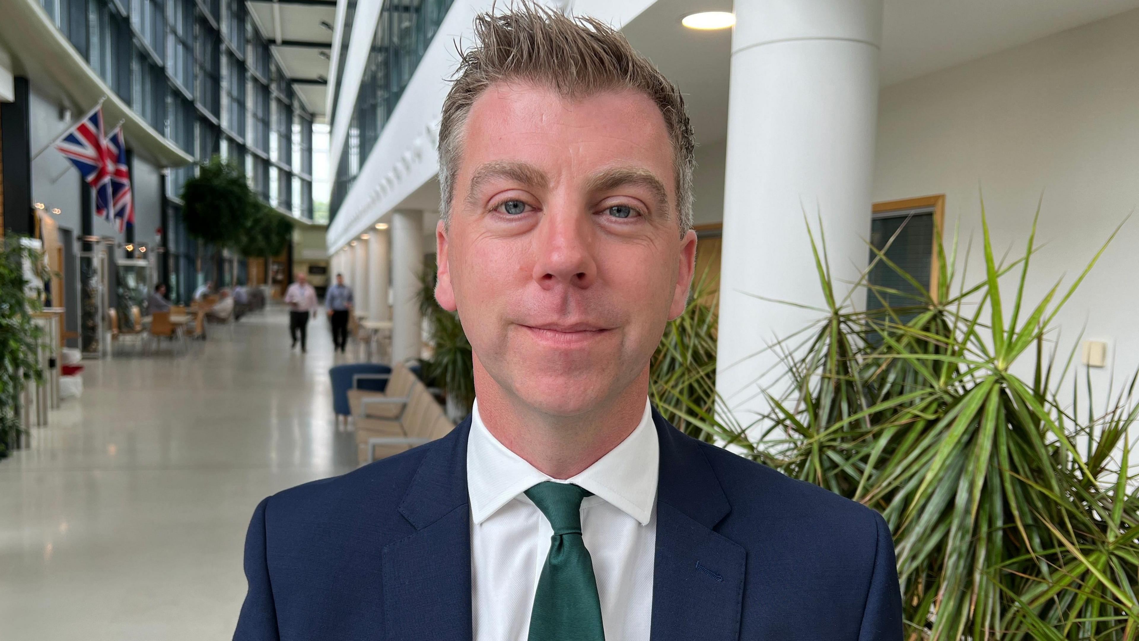 Deputy Gloucestershire Police and Crime Commissioner Nick Evans. He is wearing a blue suit, a green tie and a white shirt and stood inside a building next to some plants. 