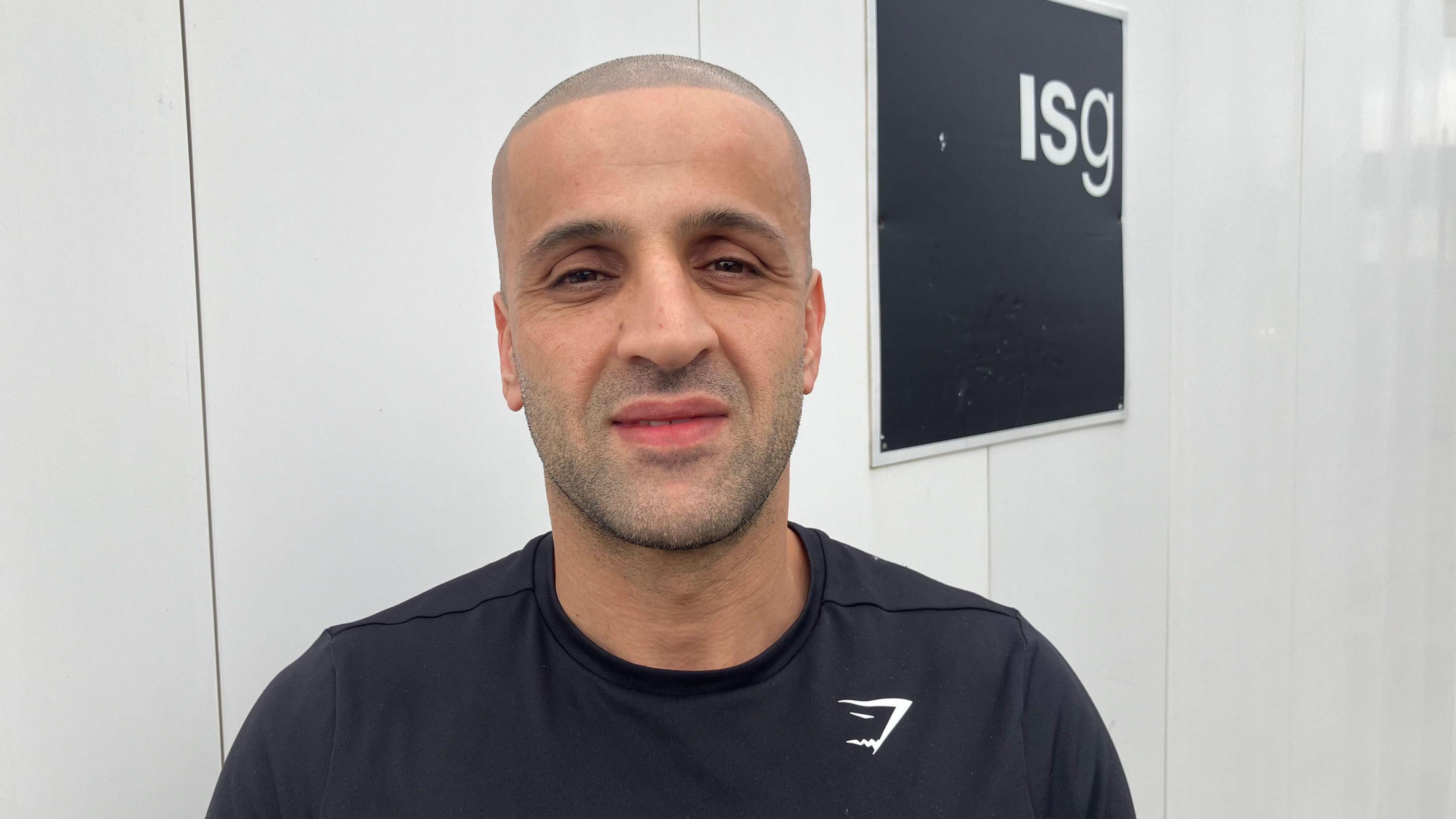 Yaz Azizi looking at the camera smiling. He is wearing a black top with a small white logo on right. He is stood in front of white boards which has a black sign on it which has the letters 'ISG' on it.