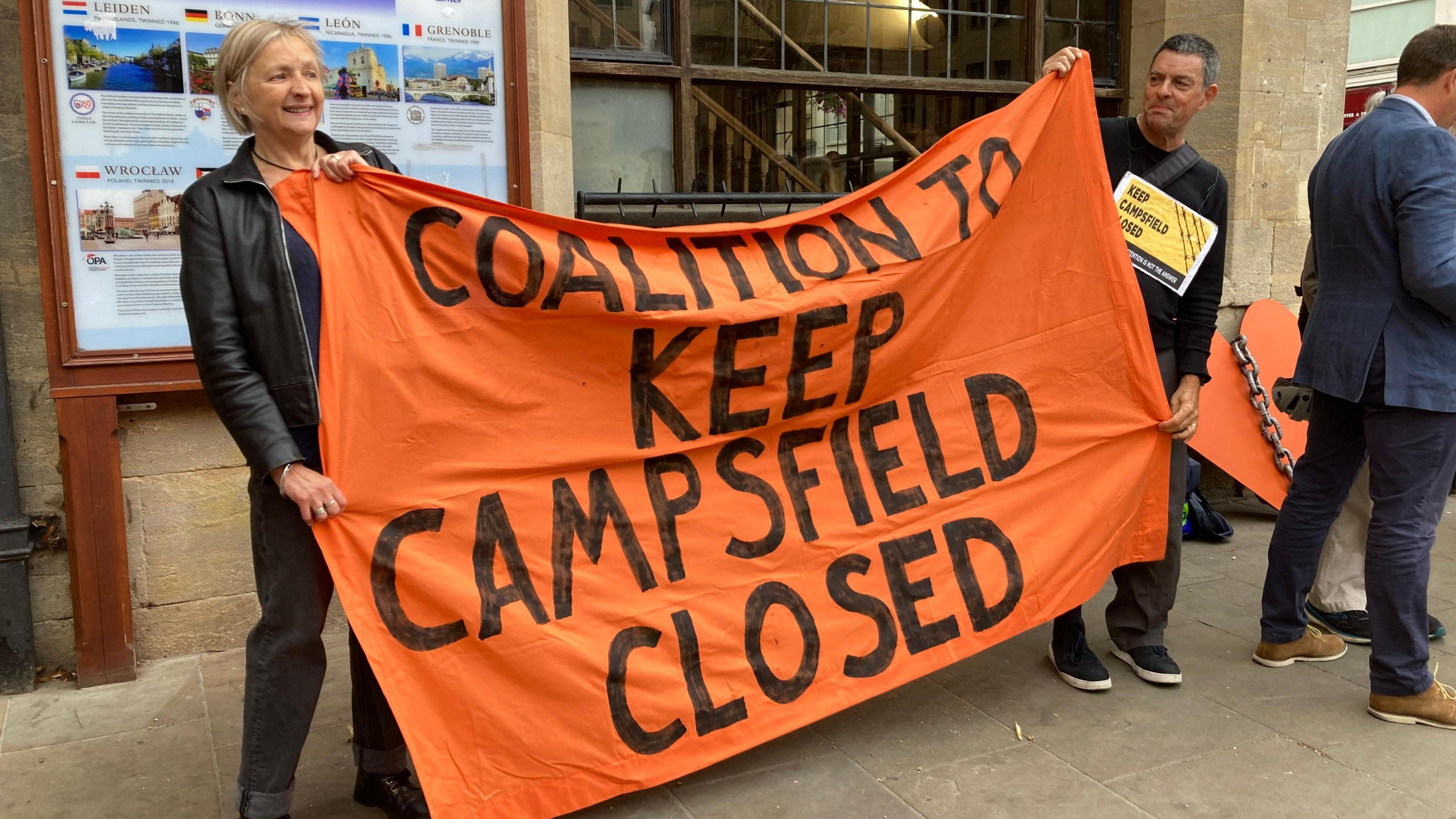 Two people holding a large, bright orange banner with "Coalition To Keep Campsfield Closed" written on it, in front of a sandstone building