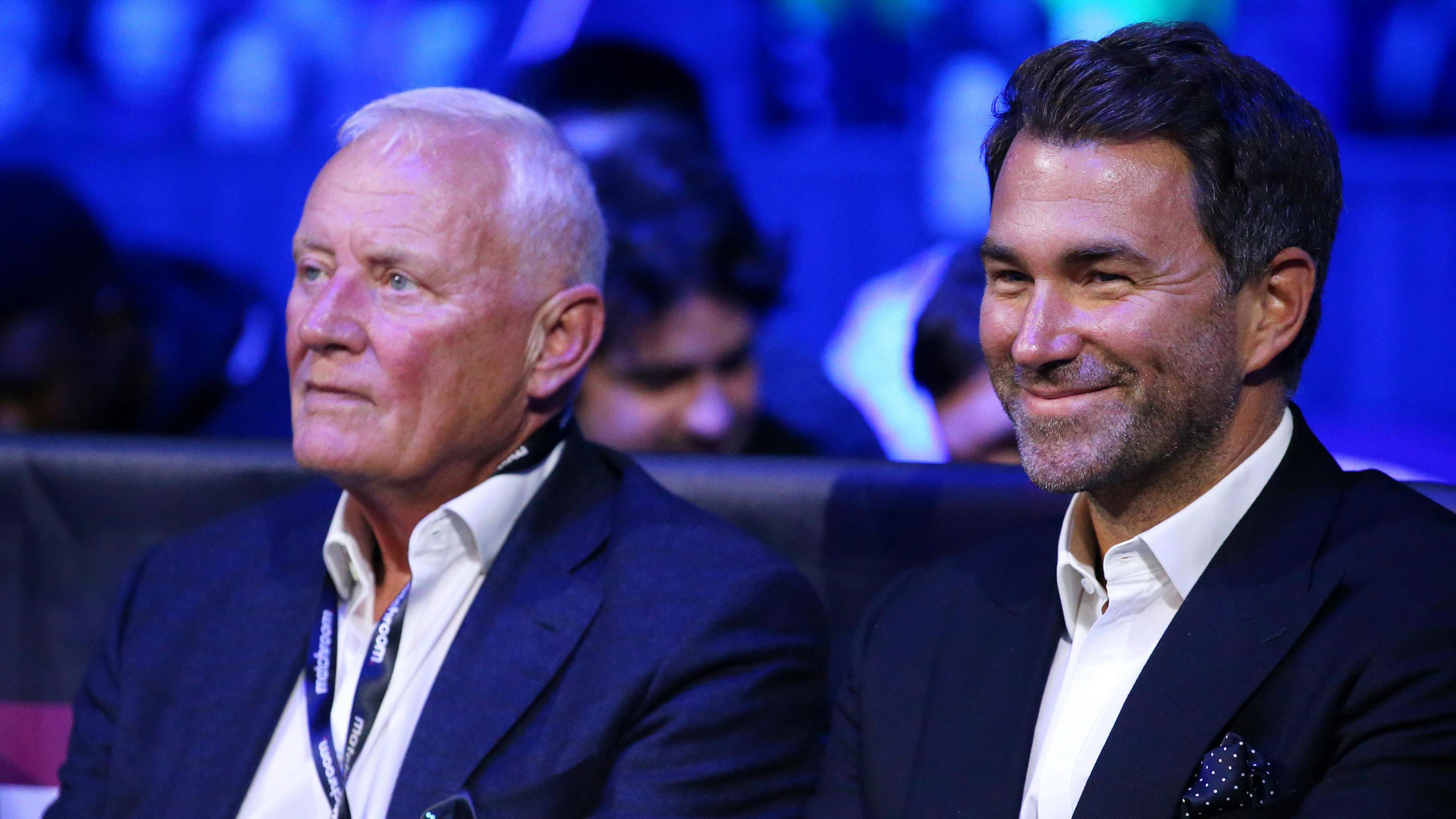 (L-R) Barry Hearn is wearing a white shirt and a navy blue blazer. His son, Eddie has dark brown hair and is smiling while sitting next to his dad. He is wearing a white shirt and a black blazer.