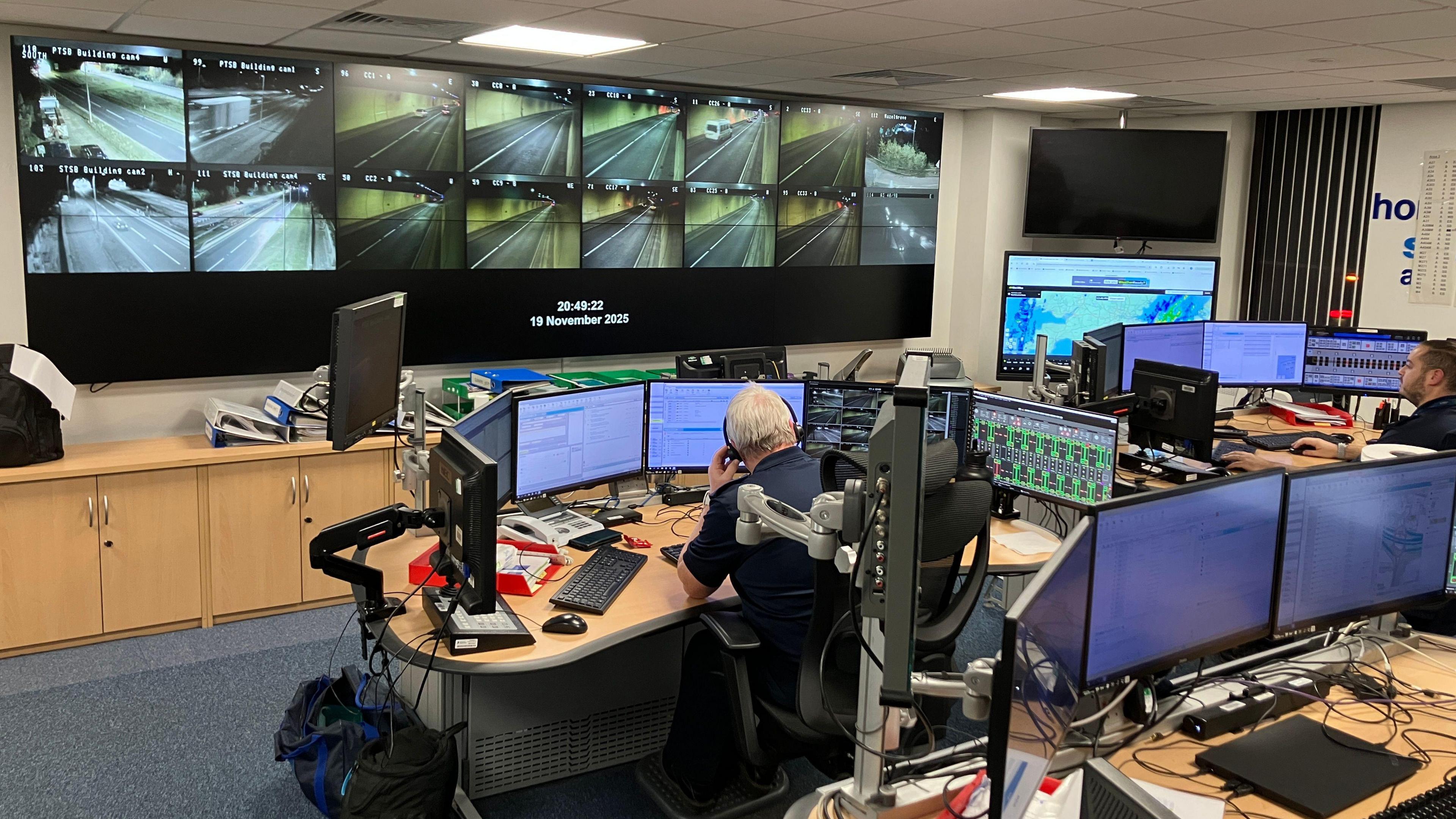 A control room in a tunnel, showing about a dozen screens on a wall with four large desks facing towards them. Two people are sat at the front two desks