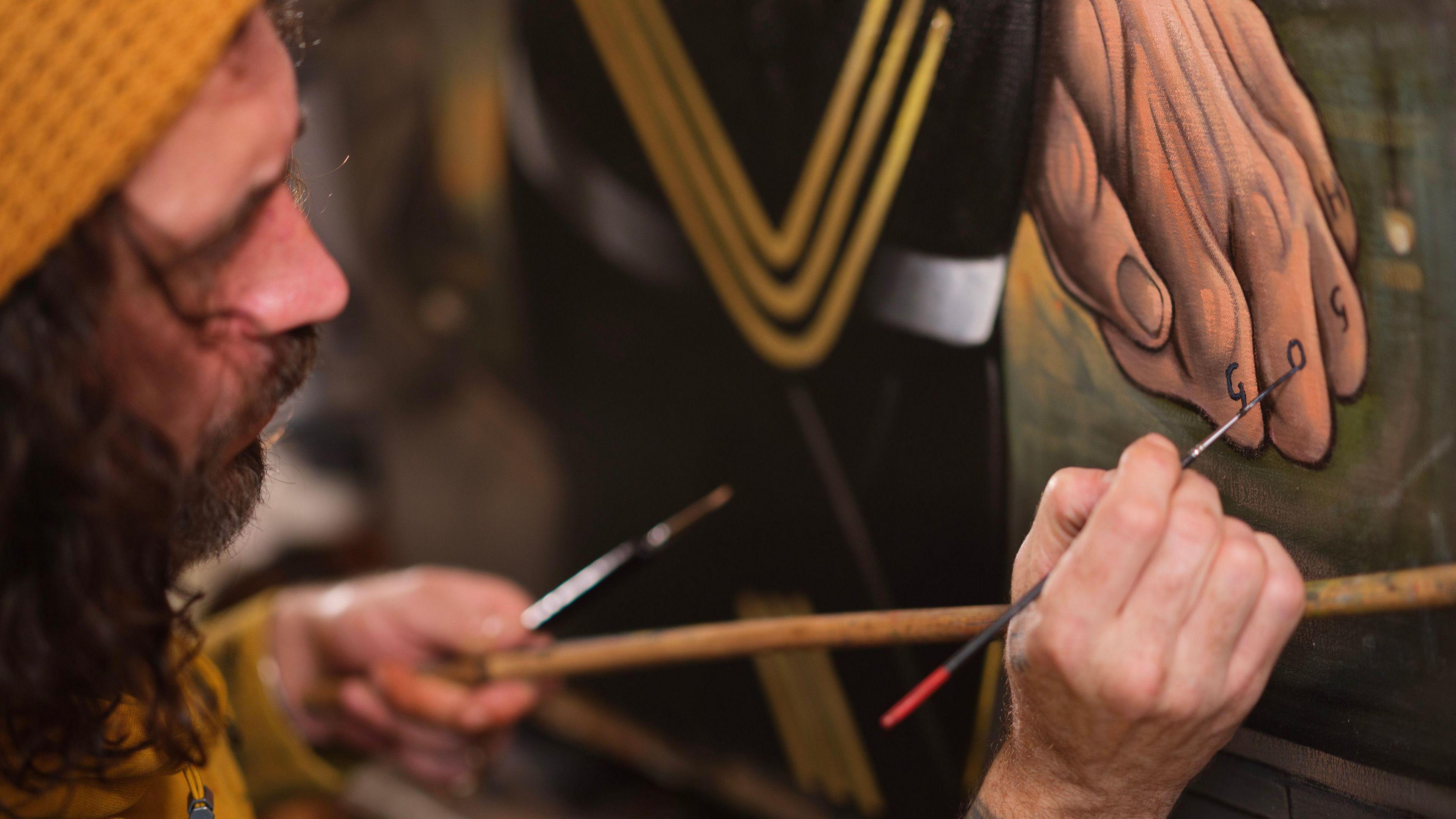 A close-up of Ross Muir who has shoulder-length, curly hair and is wearing a yellow beanie painting GOGH on the knuckles of a portrait.