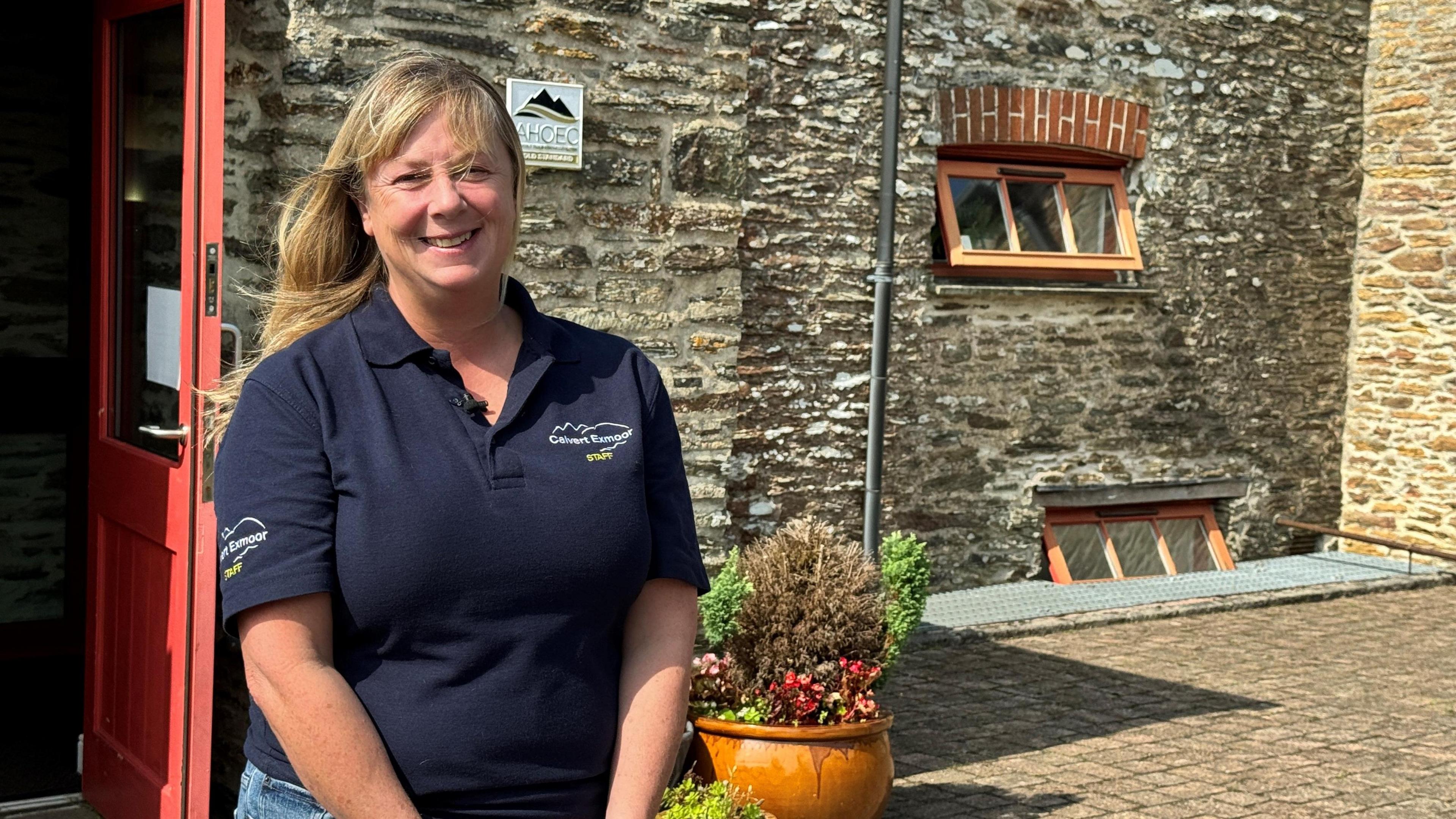 Jenny Young wearing a Calvert Exmoor T-shirt is standing outside the front door of the activity centre smiling at the camera