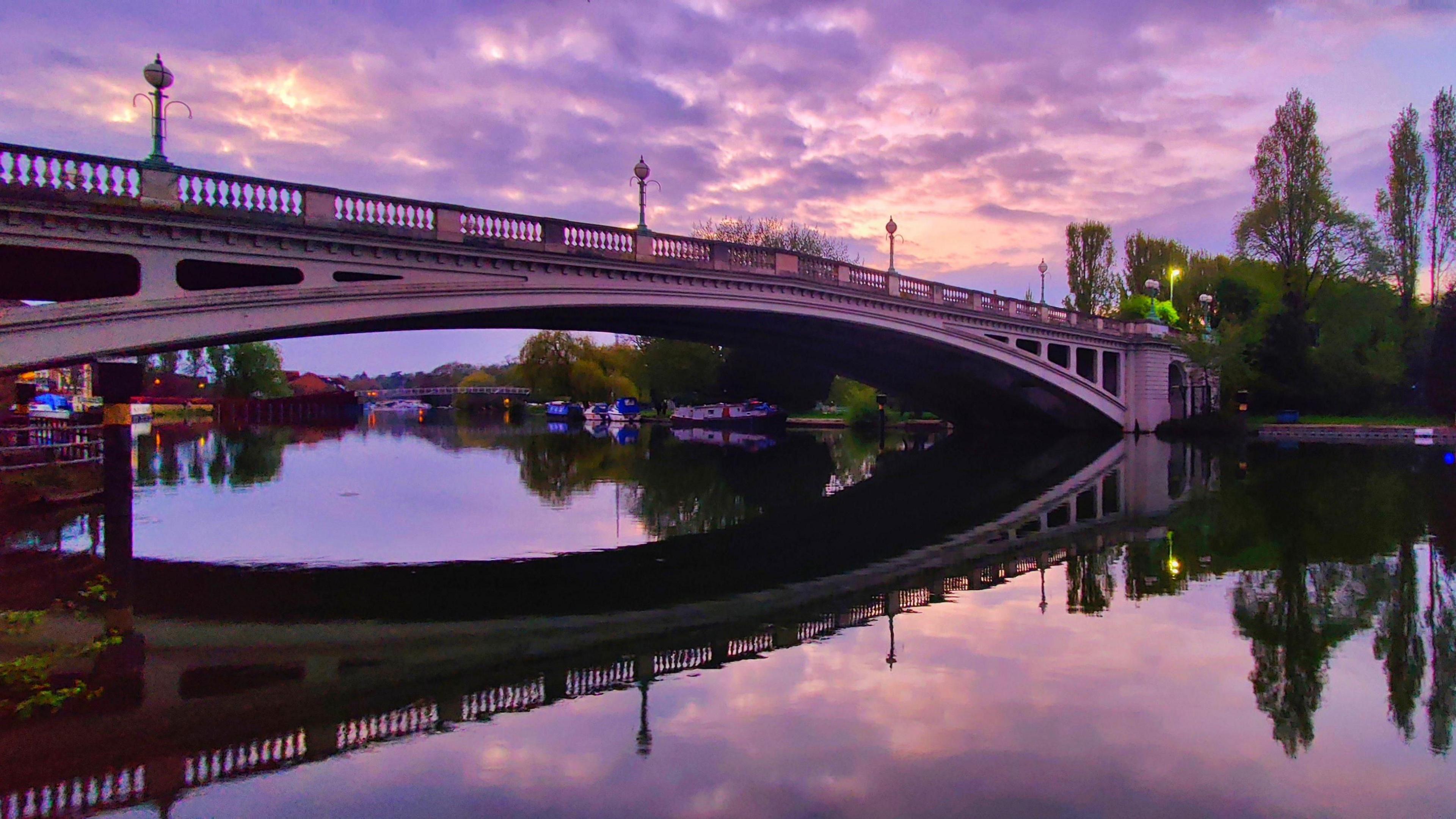 A beautiful purple sky over Caversham Bridge in Reading. The sky at sunset is lighting up the clouds over a stone bridge. The purple colour is reflected in the clouds and the river. At one end of the bridge you can see green trees. 