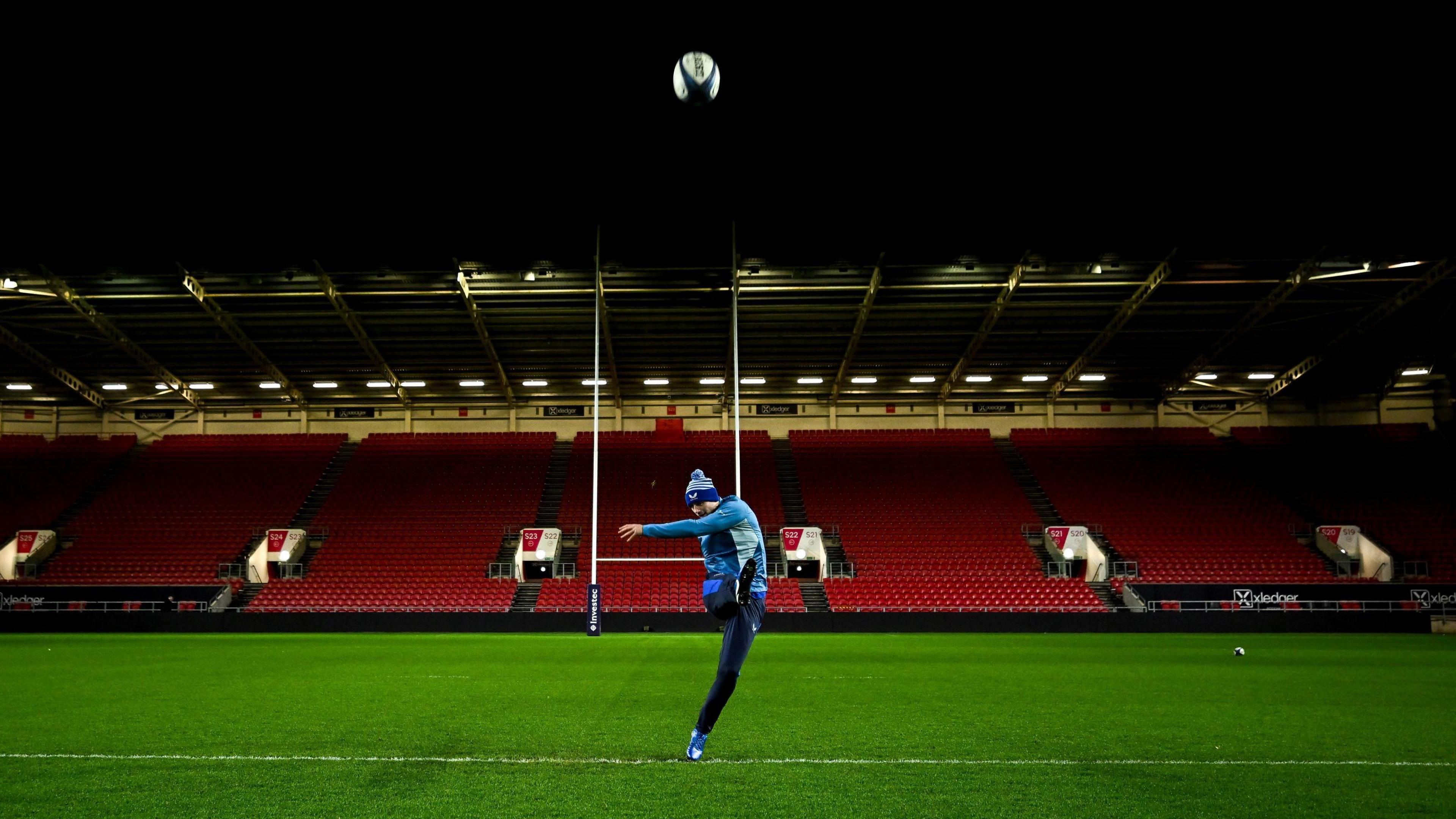 Ross Byrne of Leinster practice his kicking as the players from his team train on the pitch at Ashton Gate ahead of their game with Bristol. The stadium lights are on but the stands are empty behind him as he sends the ball through the air in the direction of the camera. He is wearing a training kit that is a mixture of light and dark blue, including a blue bobble hat