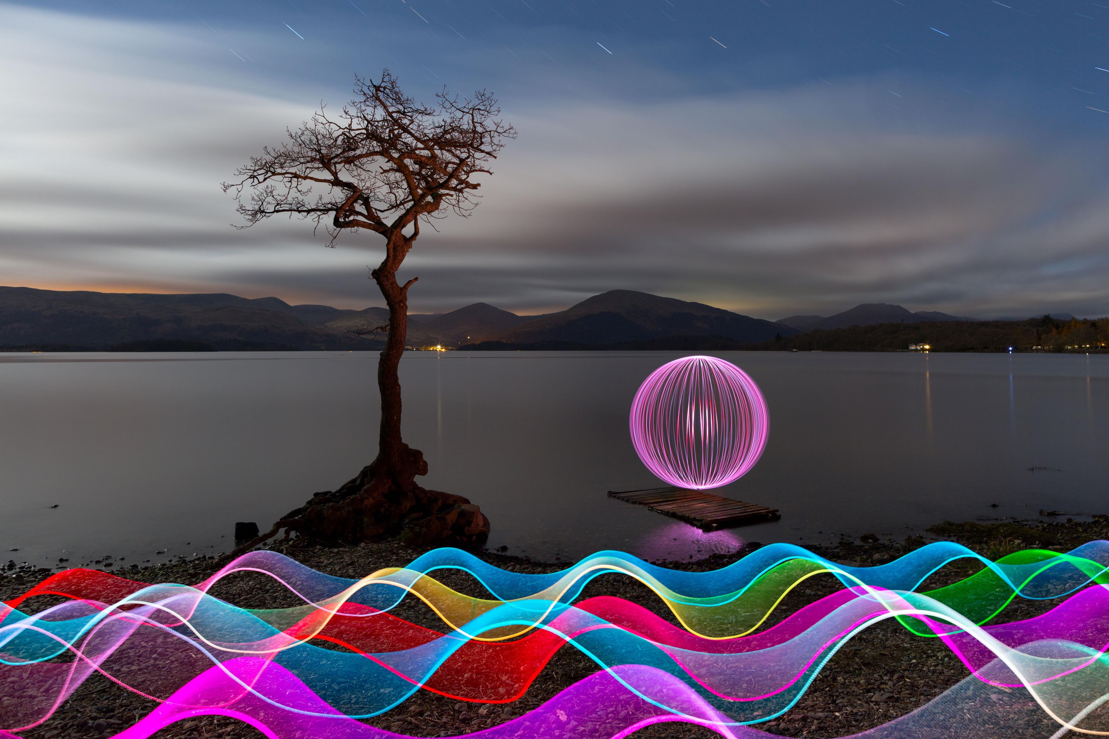 Ribbons of rainbow light are at the forefront of the image along the shore. With a smaller ball of light looks like it's floating on a wooden island. There is a tree illuminated by the background. 