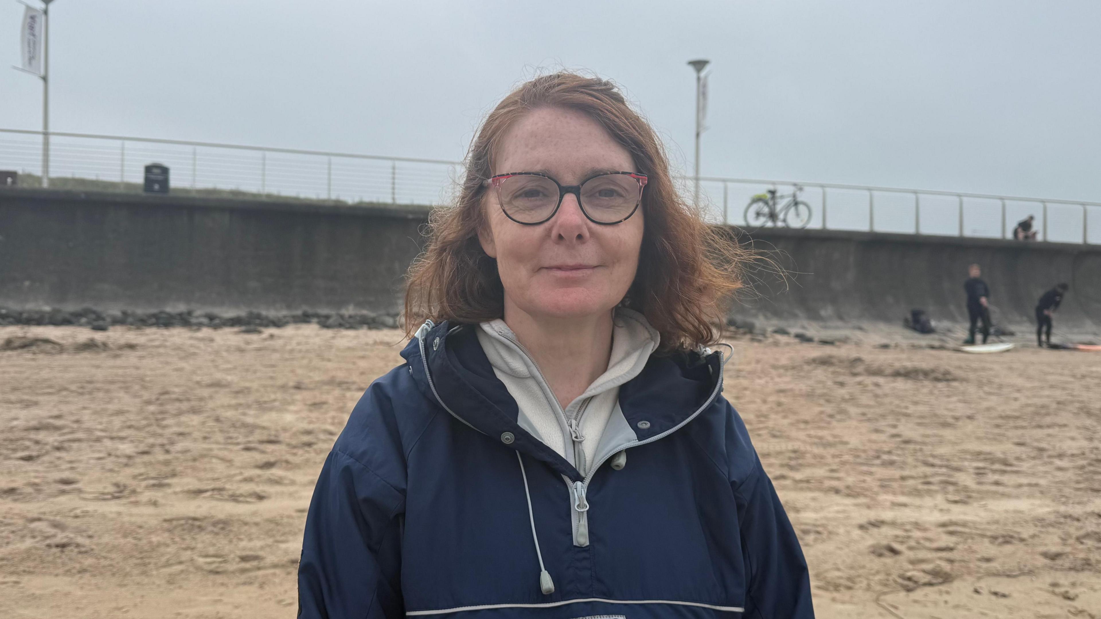 Norma Rae standing on the beach. She has shoulder length light brown hair, wearing glasses, a blue raincoat and grey half zip underneath.
