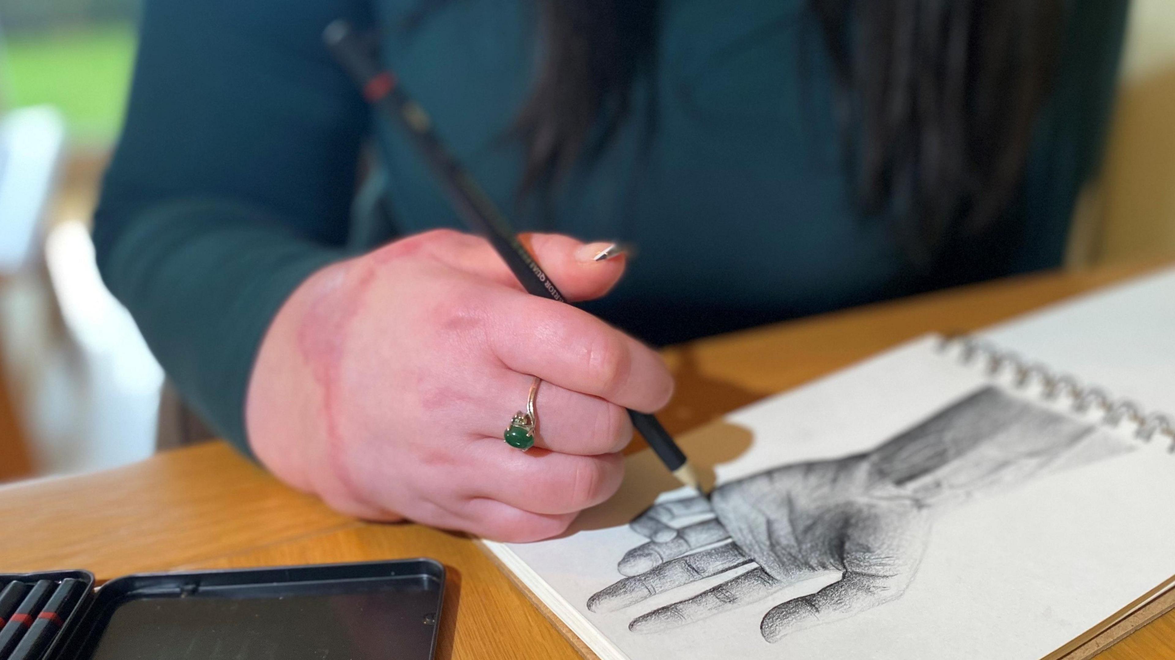 Close-up of Becca Heritage's transplanted hand holding a pencil. She is sketching an intricate picture of a hand in a sketch book. There is scarring on her hand. She is wearing a silver ring with a green gemstone and her nails are manicured.