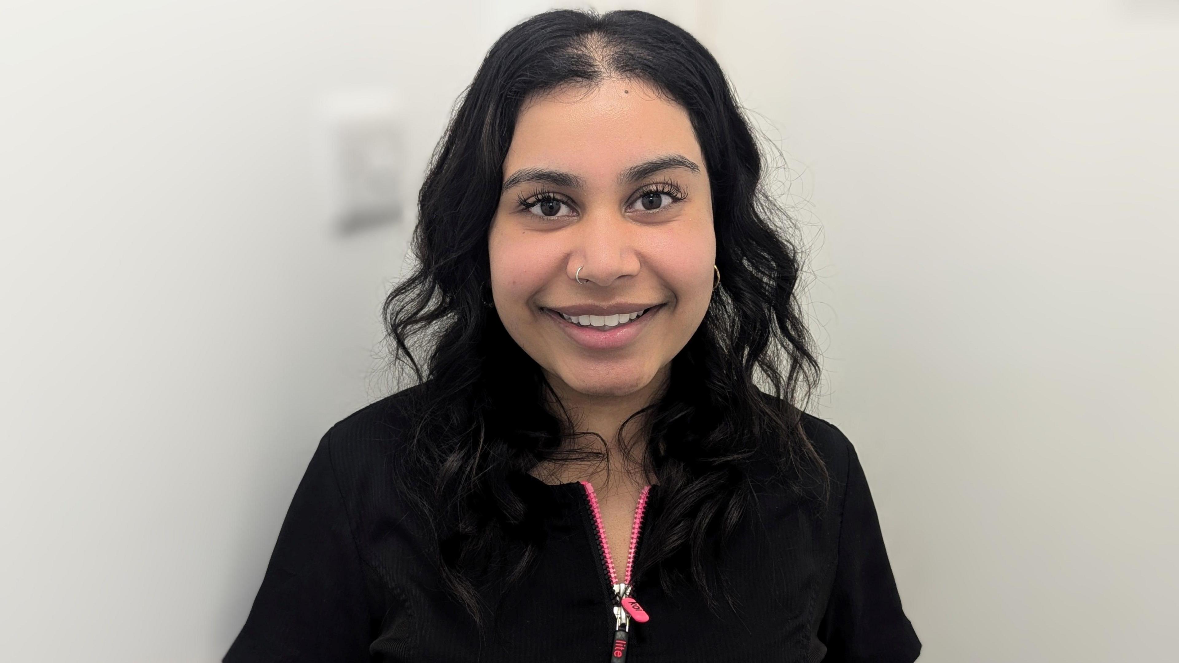 Kavita Masih - a woman with long dark hair, a nose piercing and wearing a black top with a pink zip - stands and smiles at the camera.