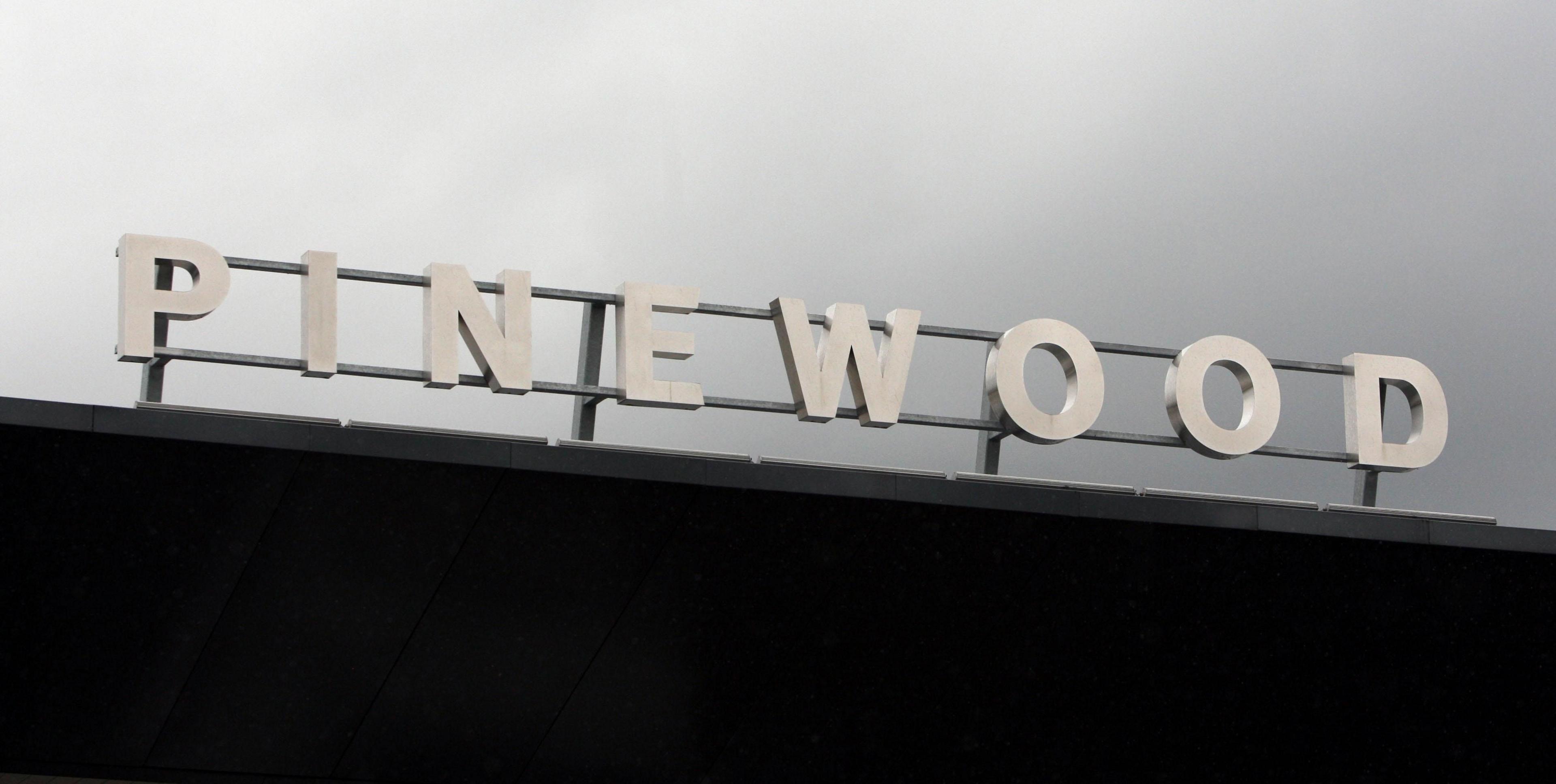 The front gate of Pinewood studios in Iver Heath. The word Pinewood has been fastened to a metal frame. They stand out against a grey sky. 