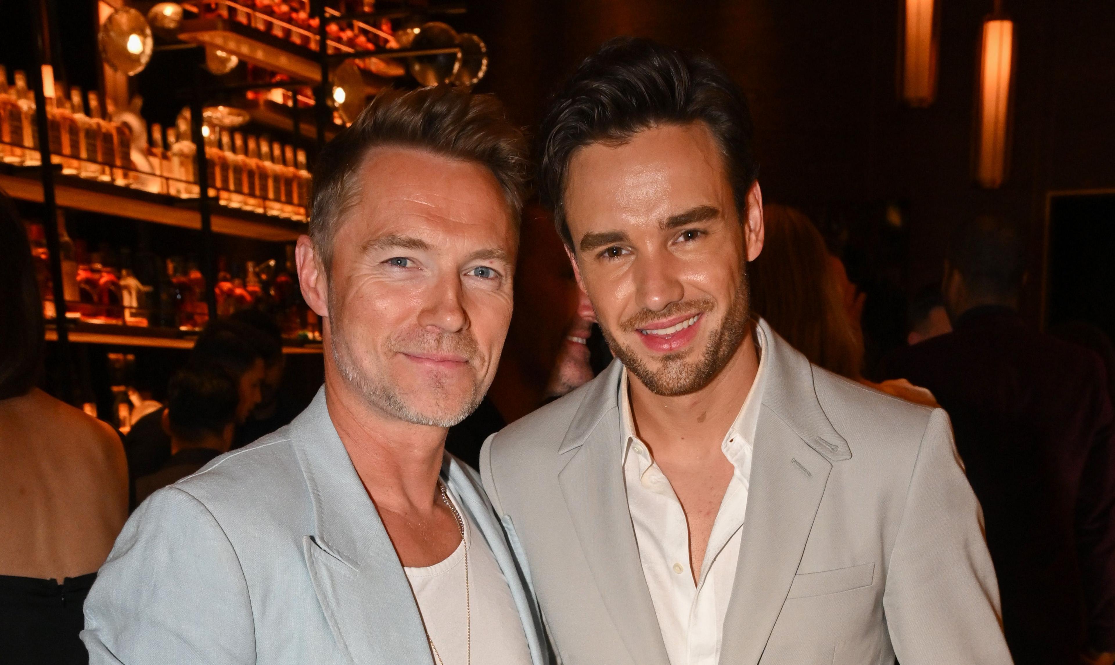 Ronan Keating and Liam Payne in a bar, both wearing light coloured suits. They are smiling at the camera.