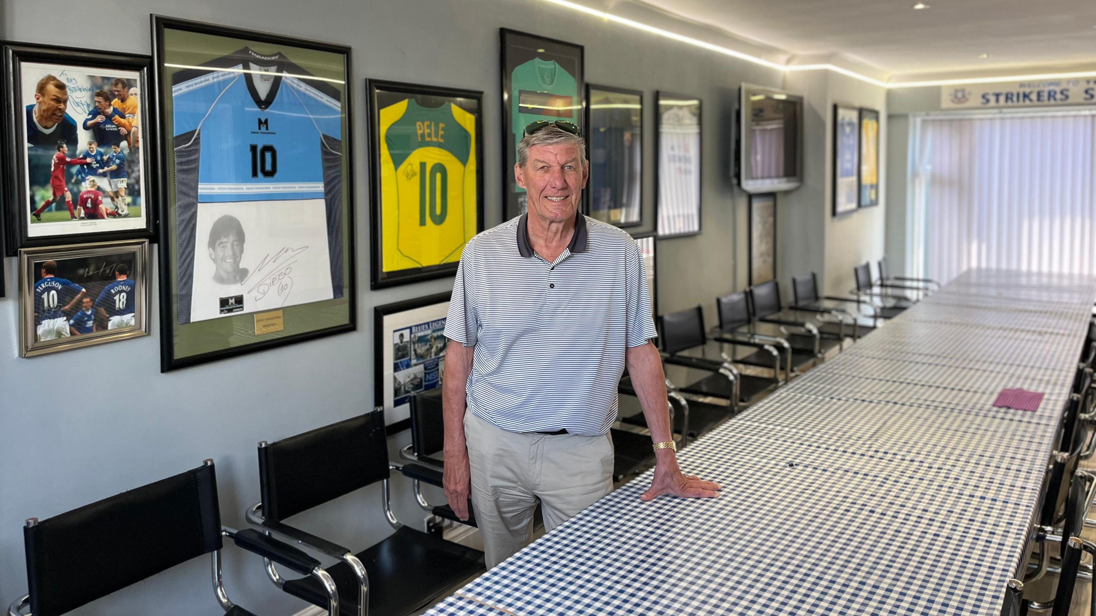 Ray Parr is leaning against a long table in the Striker's Lounge. The table is covered in a blue and white chequered table cloth and on the walls is football memorabilia including signed shirts and photographs.
