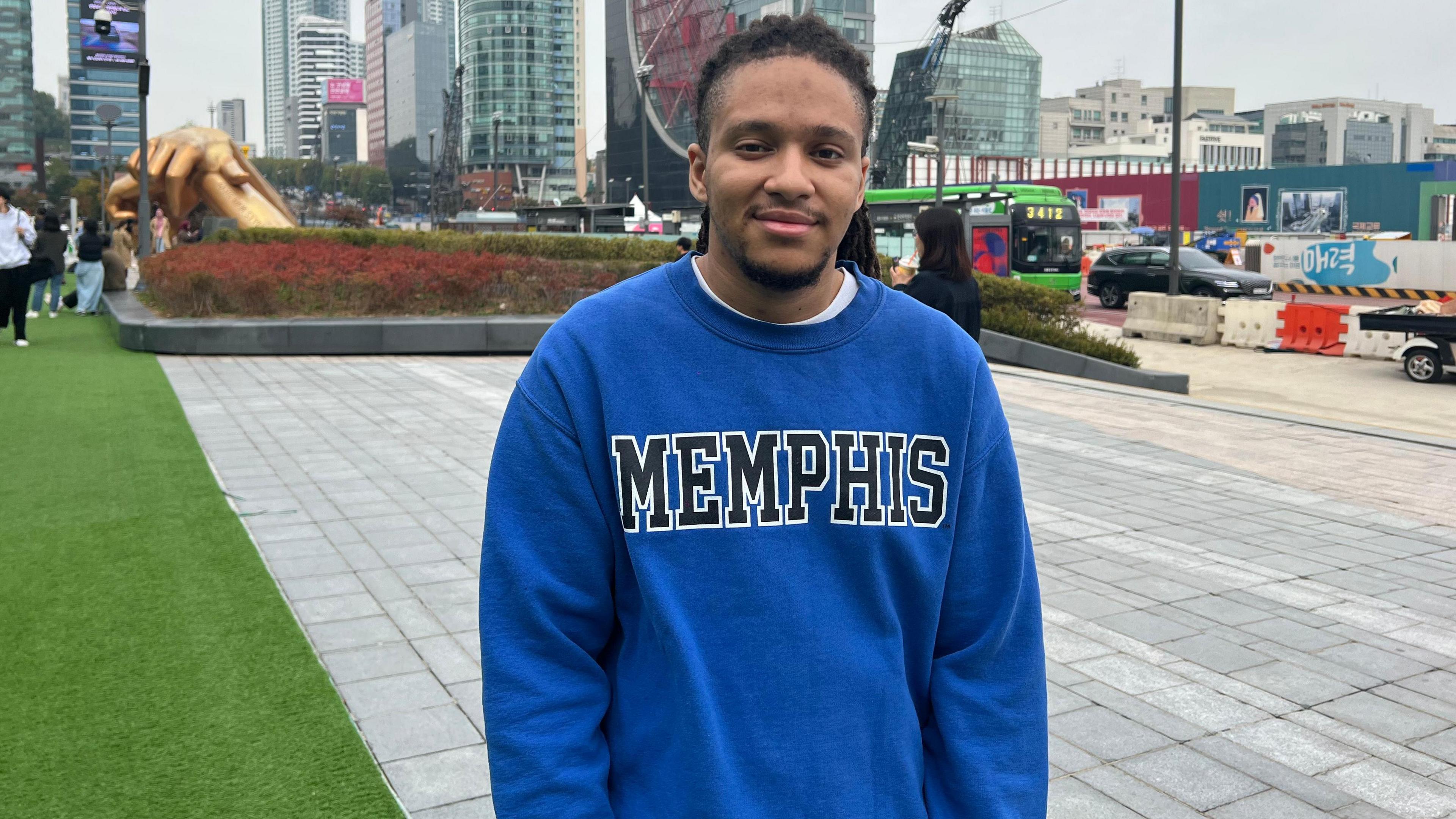 Evan Barringer is wearing a blue jumper with "Memphis" written on it. He is in a city, with towers rising behind him. On one side of the picture is a road with cars and the other a strip of grass.