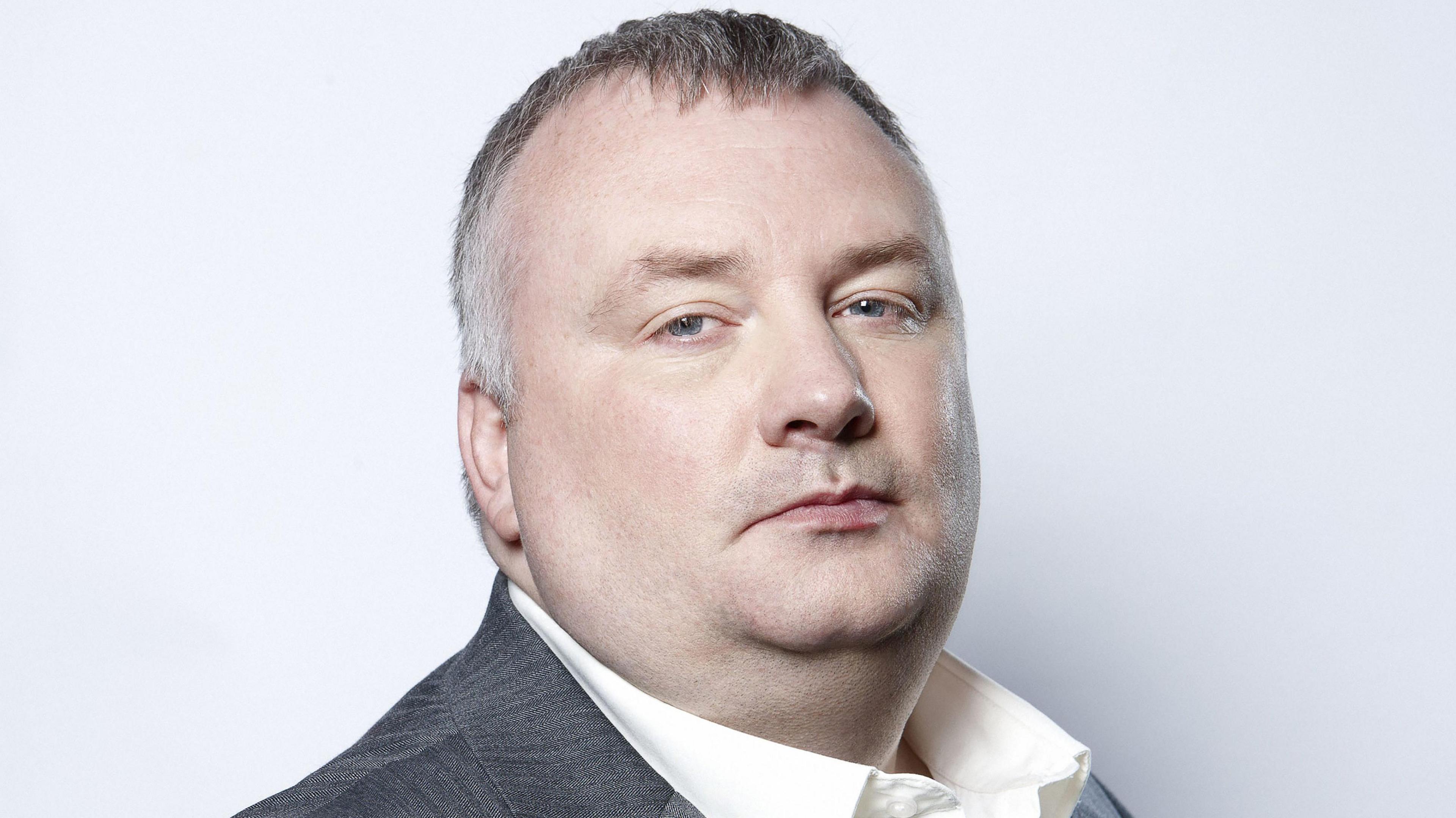 A man with short hair is turned to the side looking into the camera. He is wearing a white shirt and grey blazer.