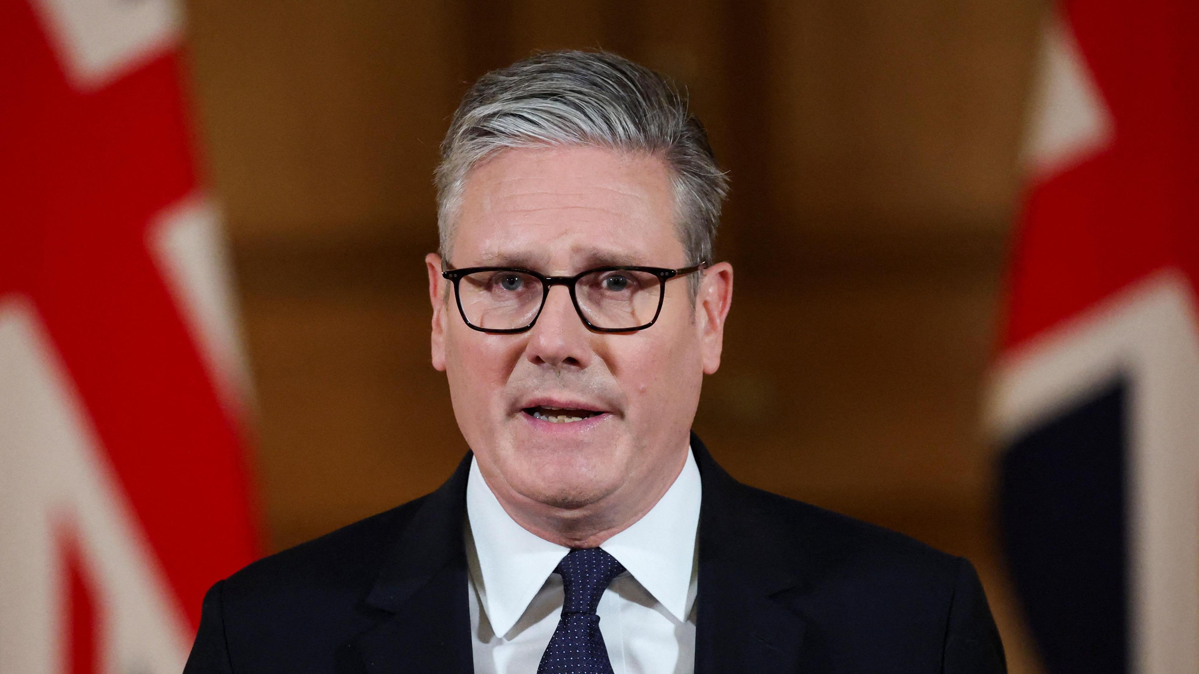 Keir Starmer. He has short grey-ish hair, wearing glasses, white shirt and dark blazer and tie. Two British flags are in the background.