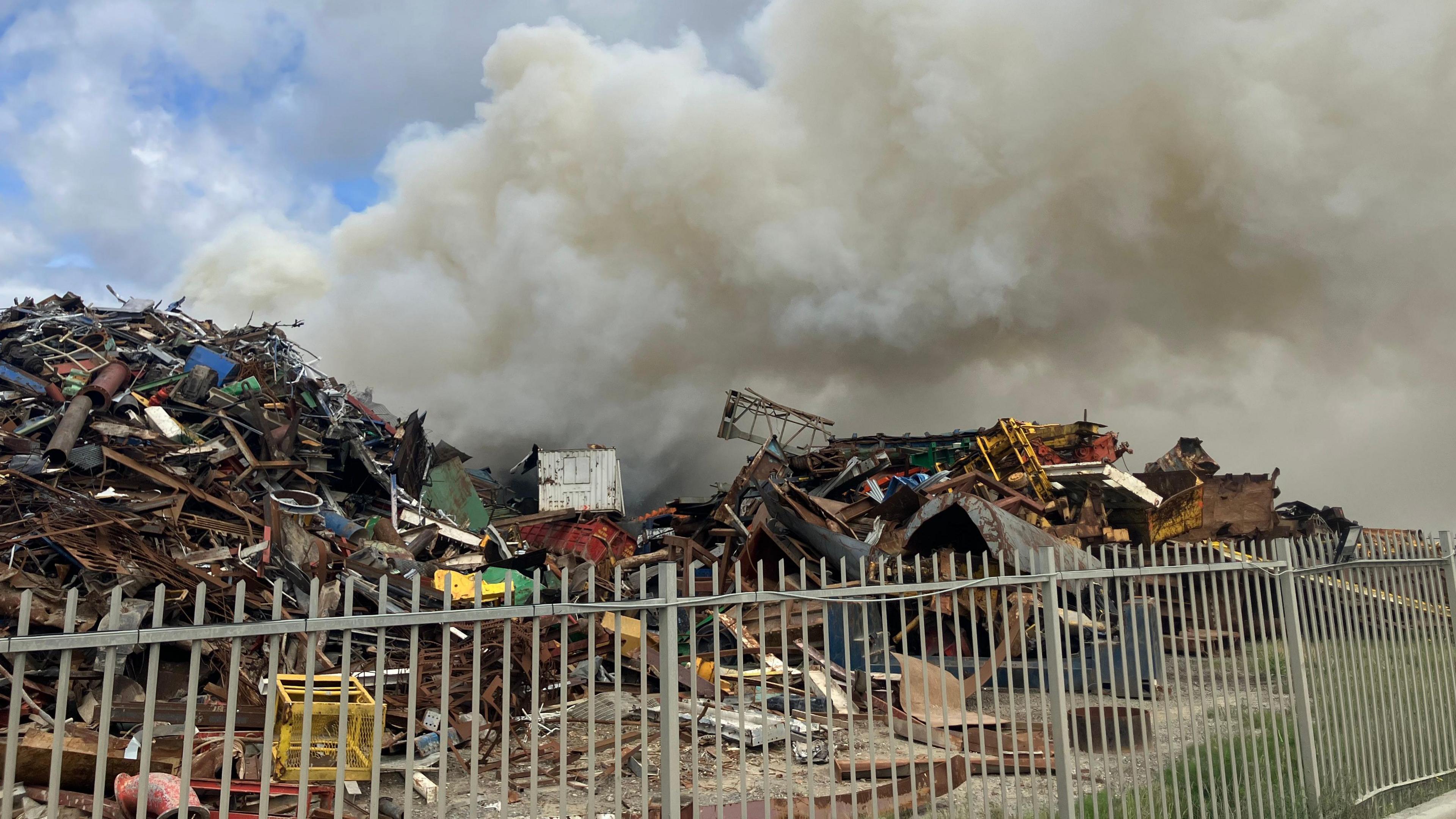 A plume of smoke billowing from a scrap yard. The grey smoke is set against a blue sky.