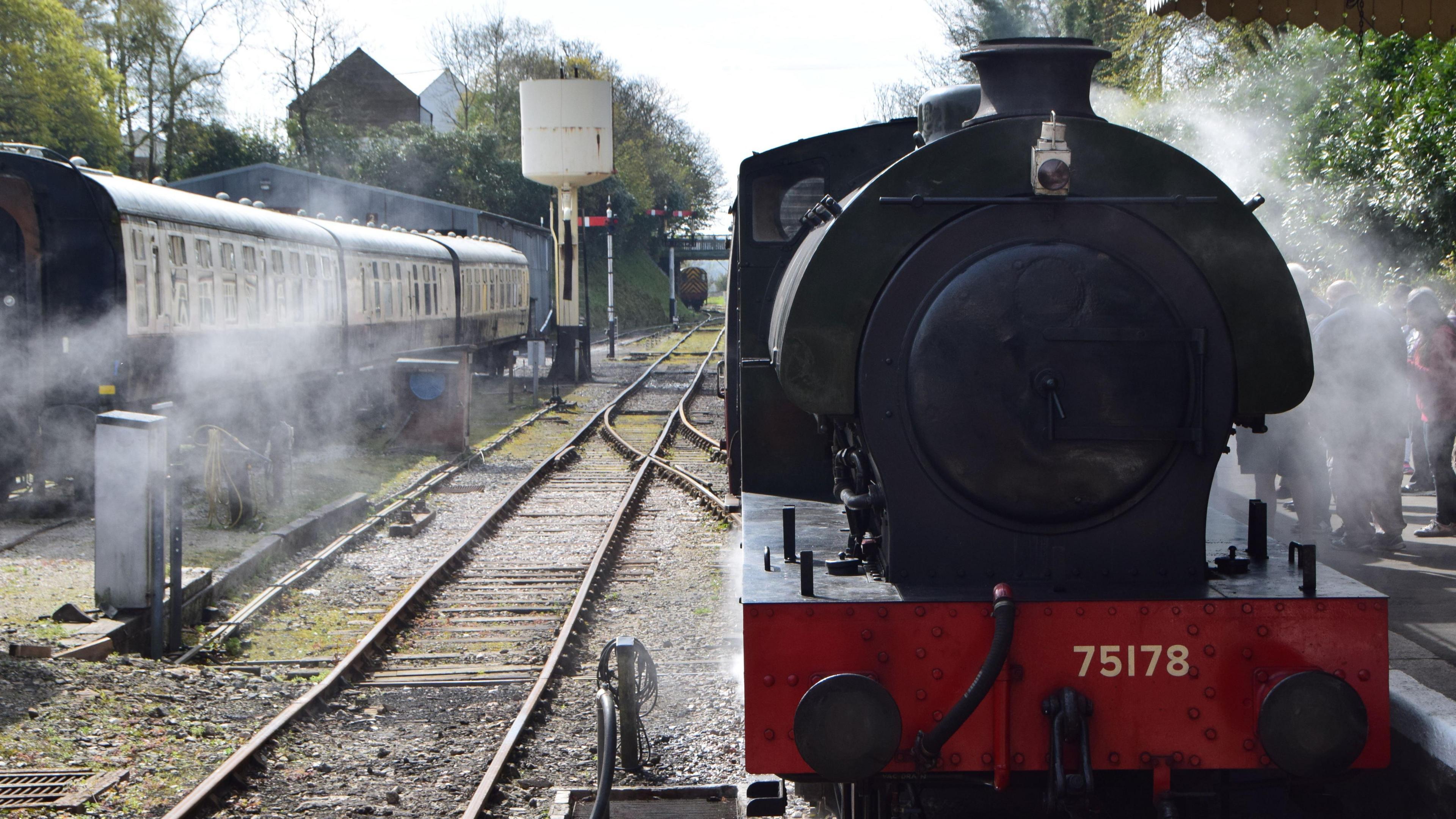 A front-facing photo of a steam engine, it's black with a red buffer and steam coming from the funnel at the top. It's on a platform and you can make out passengers waiting through the steam with a row of old fashioned carriages on the opposite side of the station.