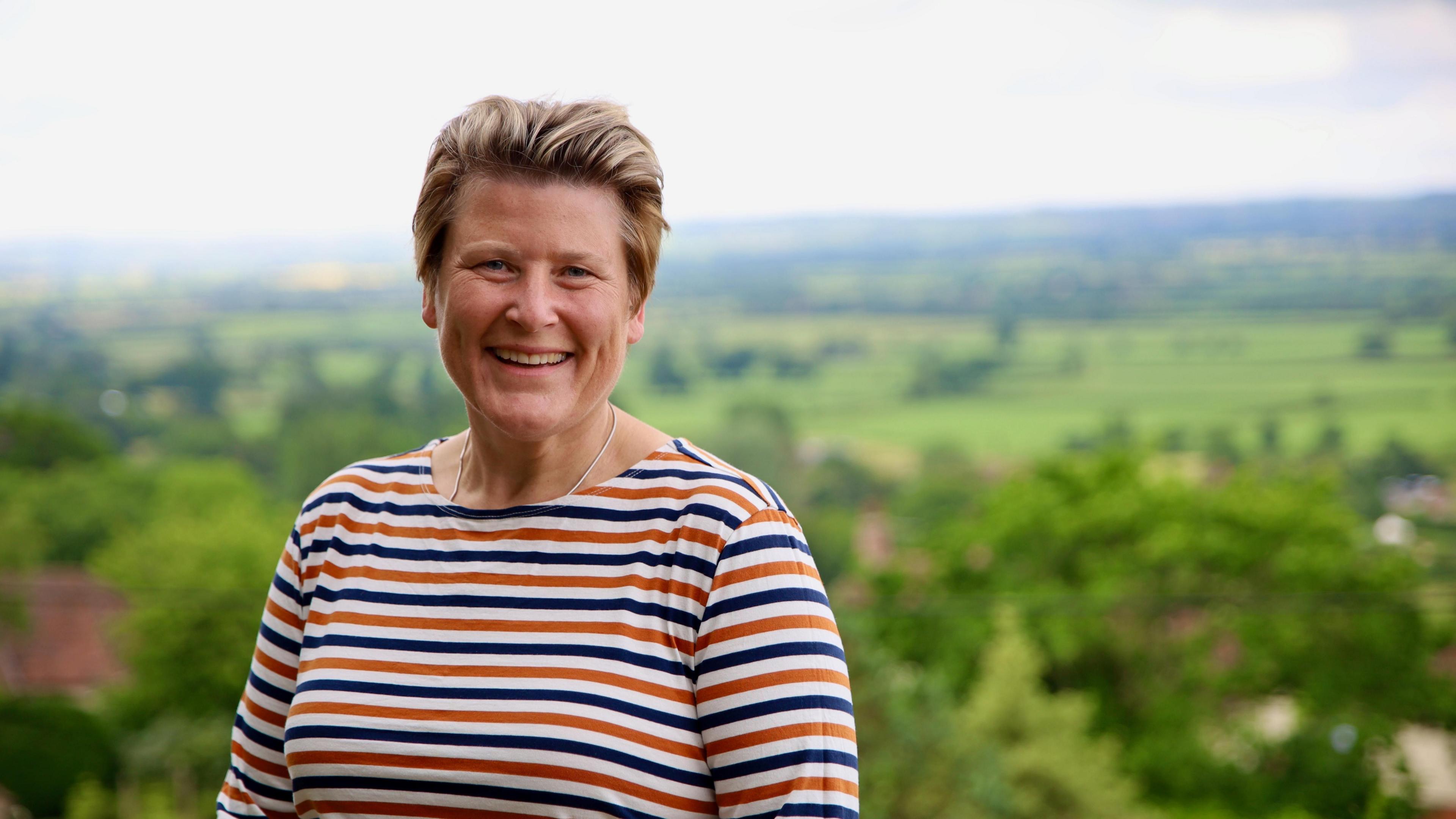 A woman in a stripy top with fields and hills behind her