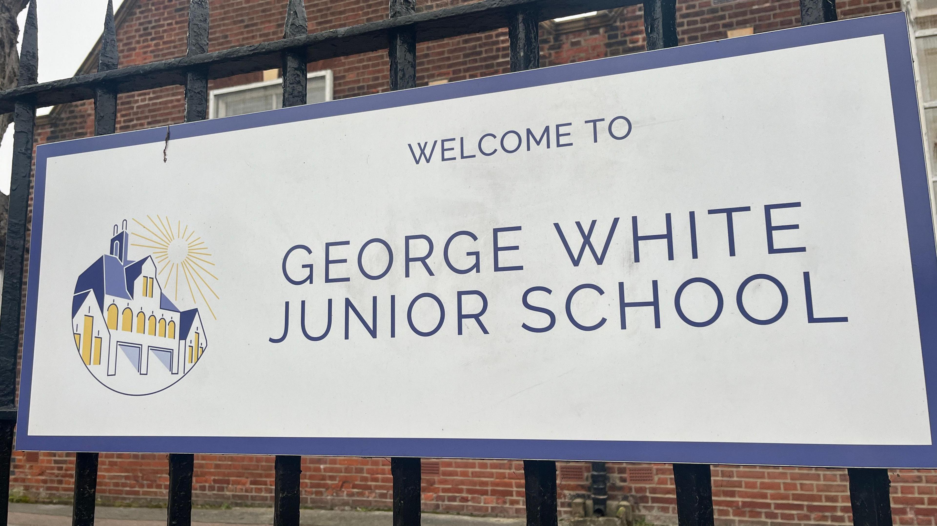 The outside of a junior school. Attached to the gates is the school sign which is white with purple writing and says WELCOME TO GEORGE WHITE JUNIOR SCHOOL.