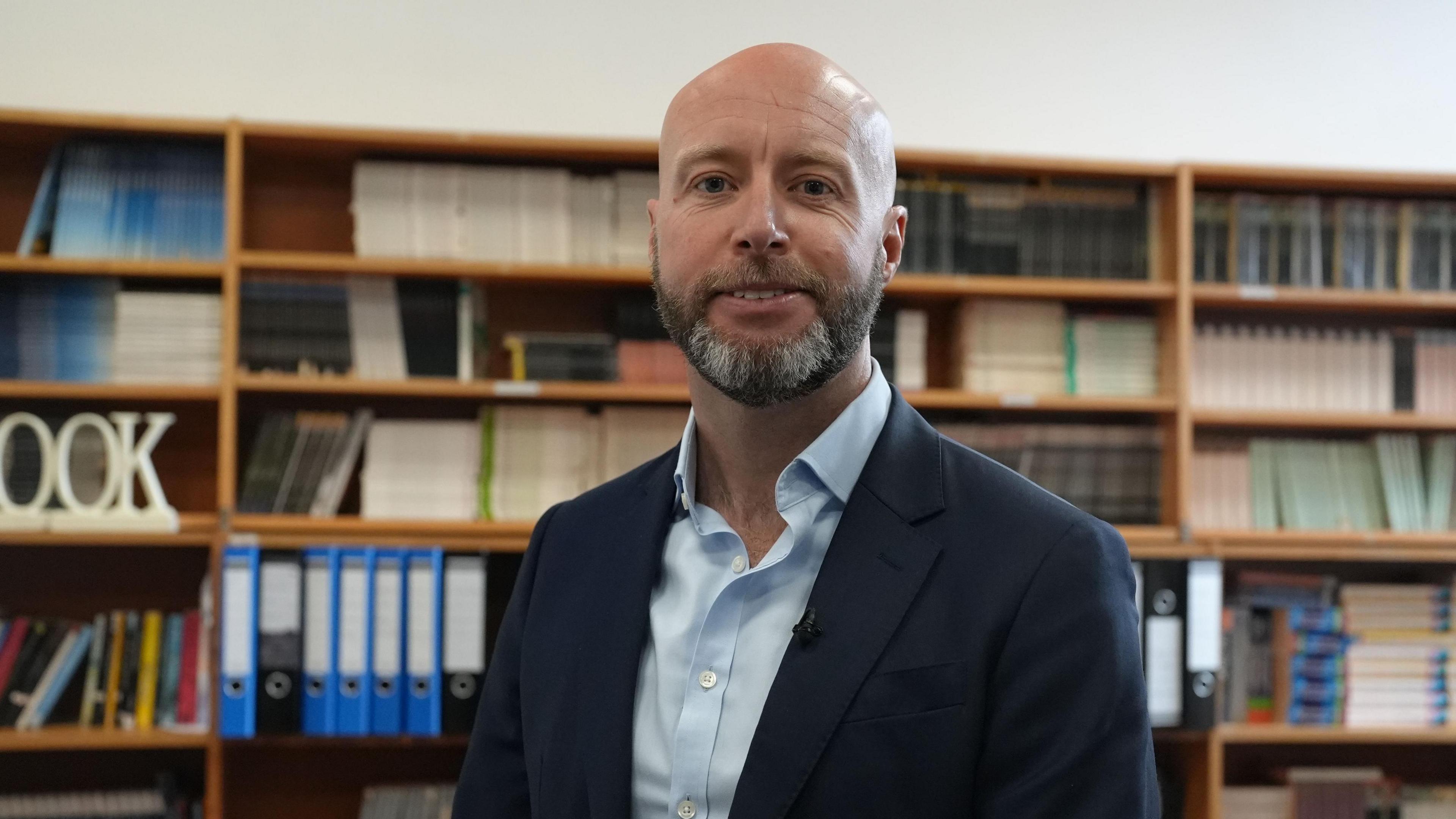 Tony Oulton looks at the camera smiling wearing a black suit jacket and light blue shirt. He has a grey and black short beard. Behind him there are shelves with rows of coloured books and folder. 