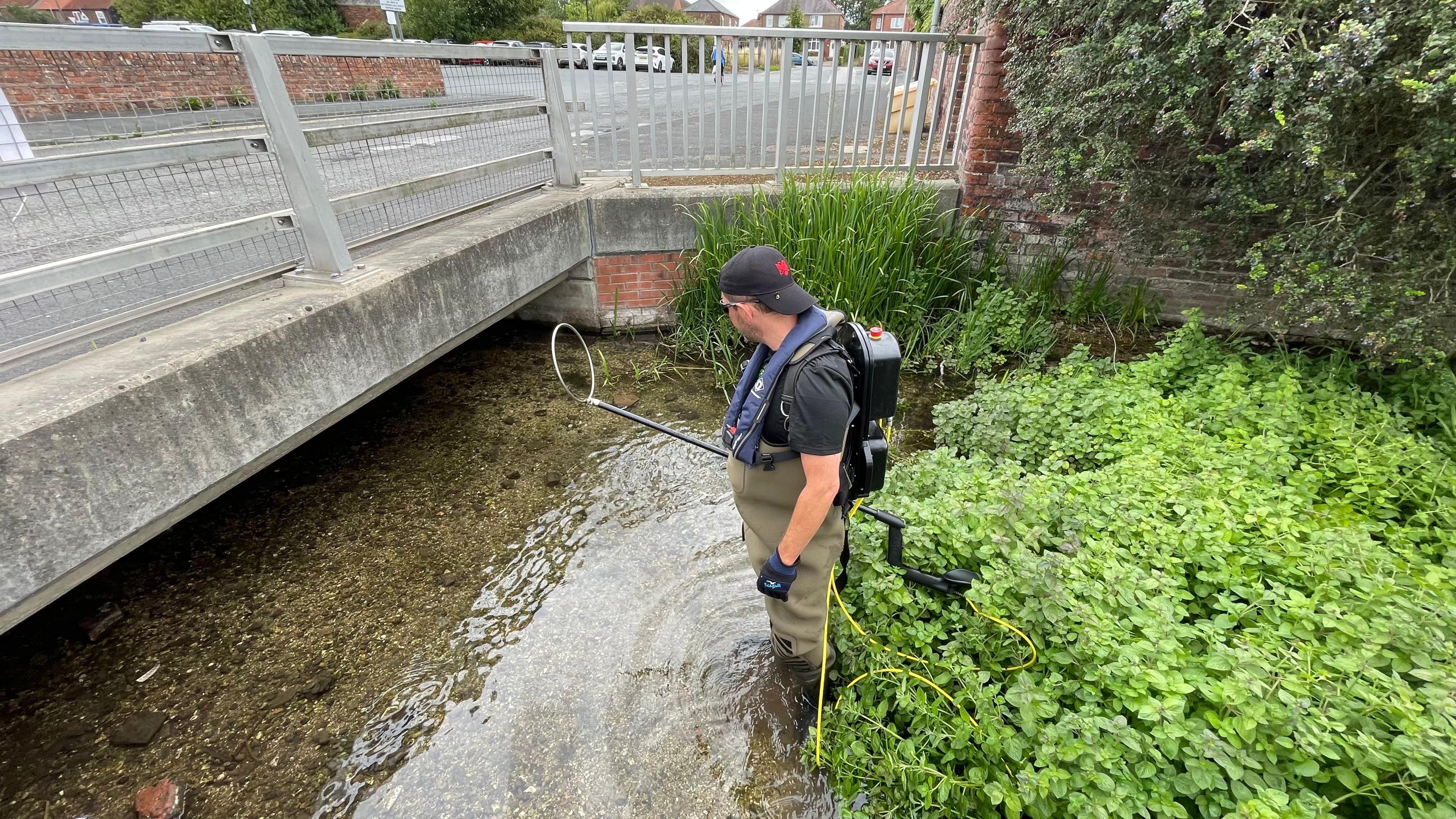 Oliver Southgate from East Yorkshire River Trust. He is standing in a stream and is carrying fishing equipment. He has a backpack on his back.