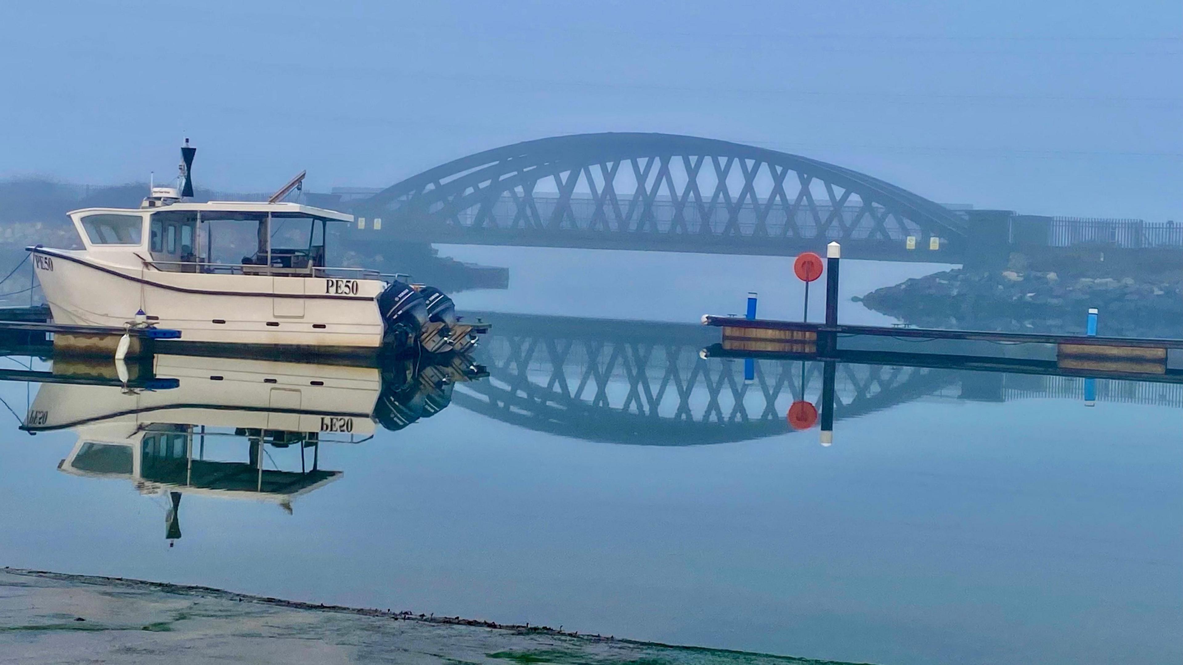 A light mist hangs over a harbour with a bridge seen in the distance and a motor boat in the foreground. The sea is calm with a light grey sky overhead.