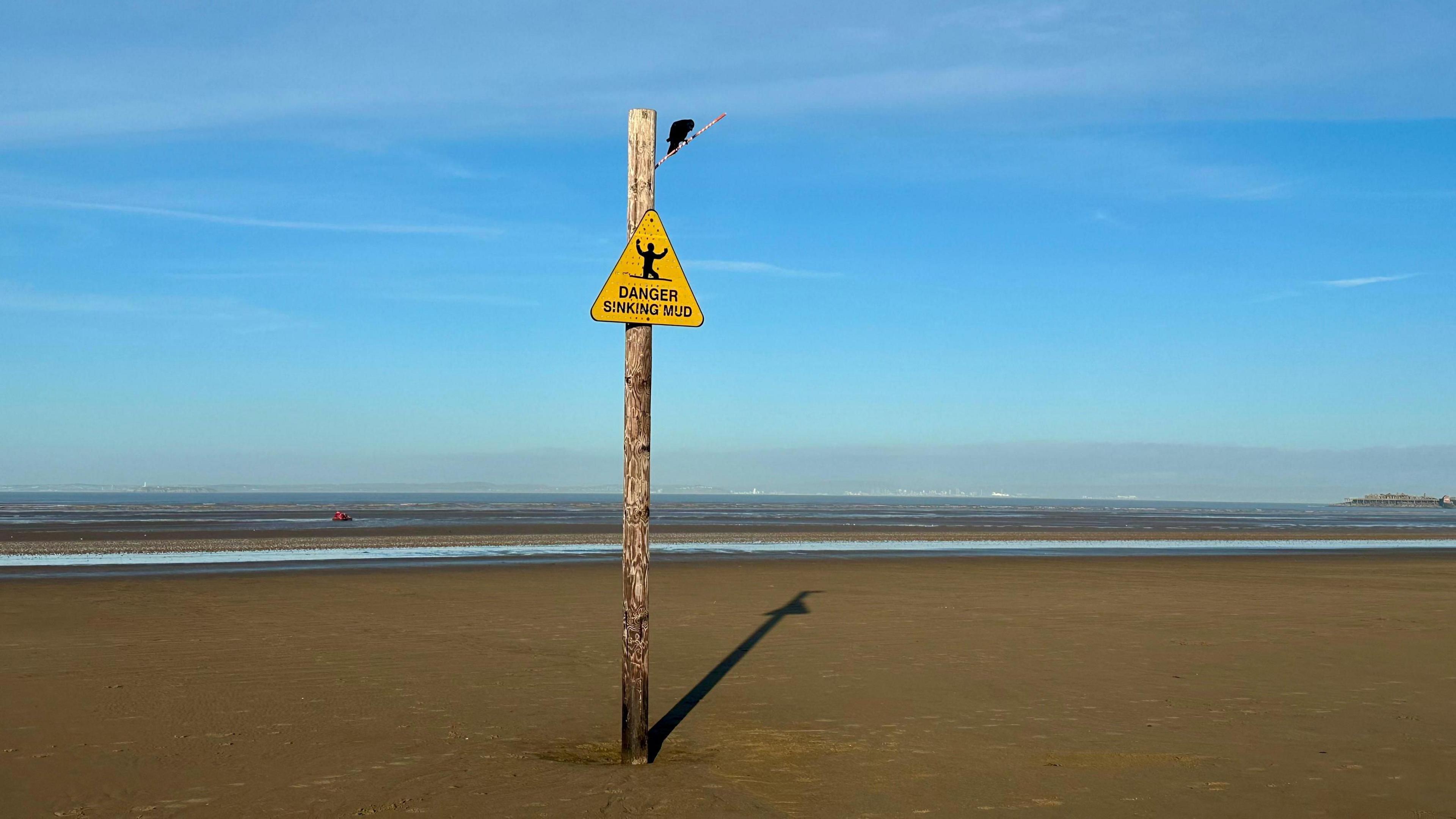 A bird is perched on a sign that reads Danger Sinking Mud. The sign is surrounded by a muddy beach with the sea at the horizon.