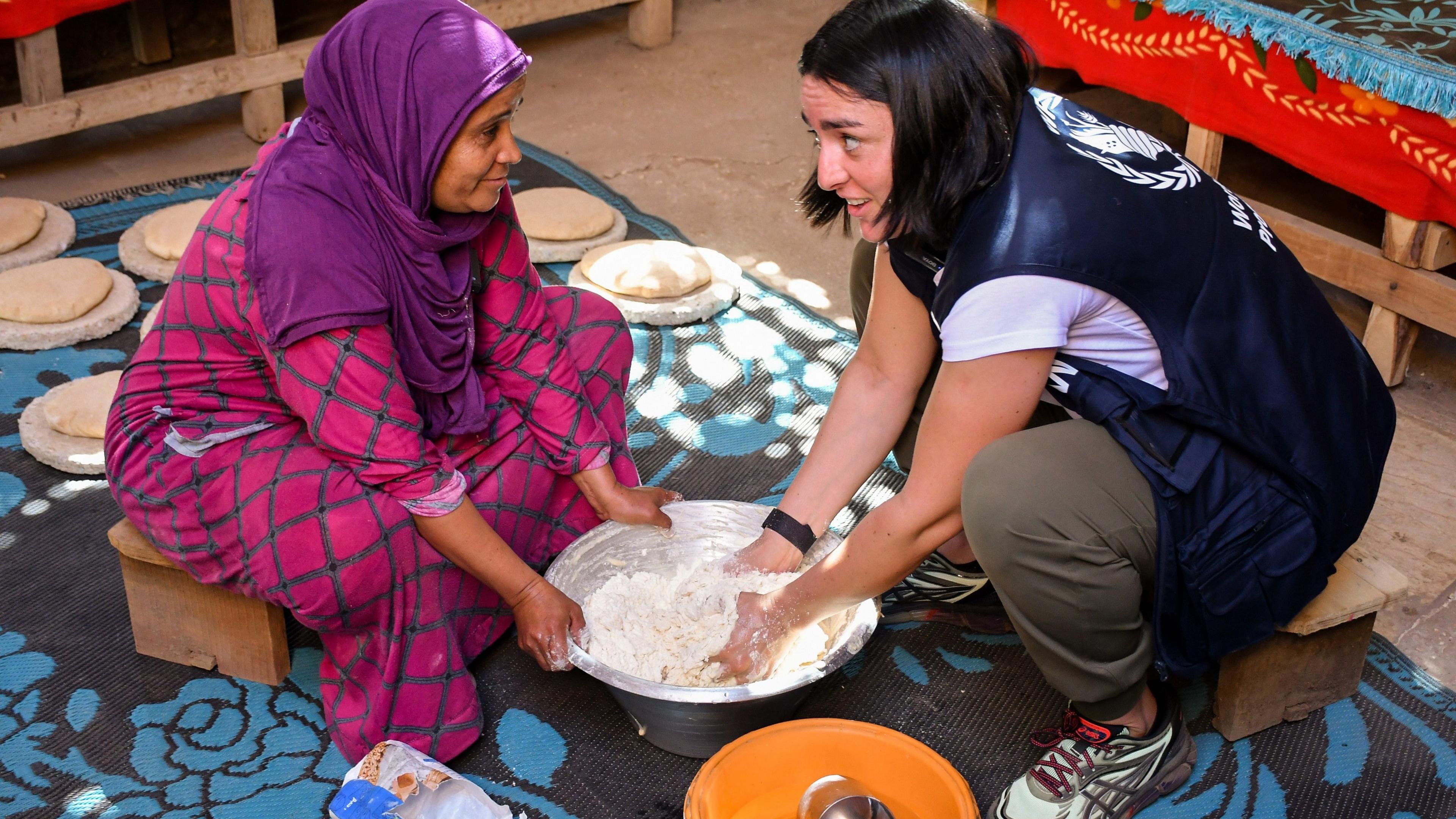 Tunisian tennis star Ons Jabeur, wearing a navy blue vest-style World Food Programme top, kneels and kneads bread in a metal bowl as she talks to a woman opposite her dressed in purple headscarf