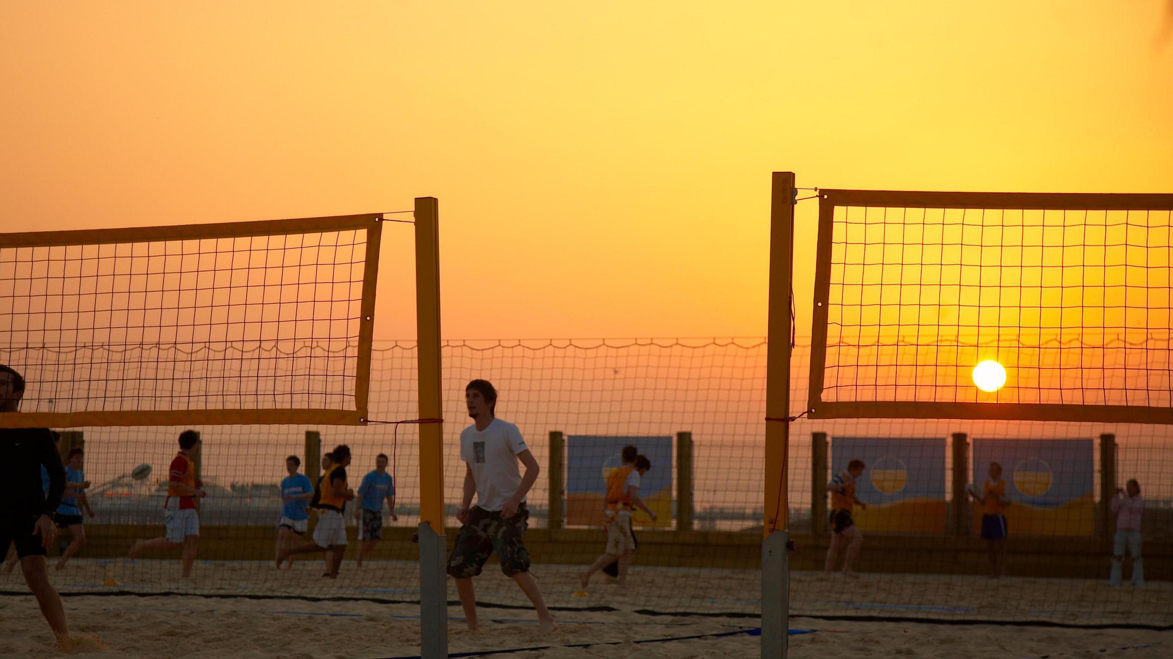 The image is of people playing beach volleyball at sunset. There are around 12 people. The sky is orange with the sun setting in the right hand side of the image. 