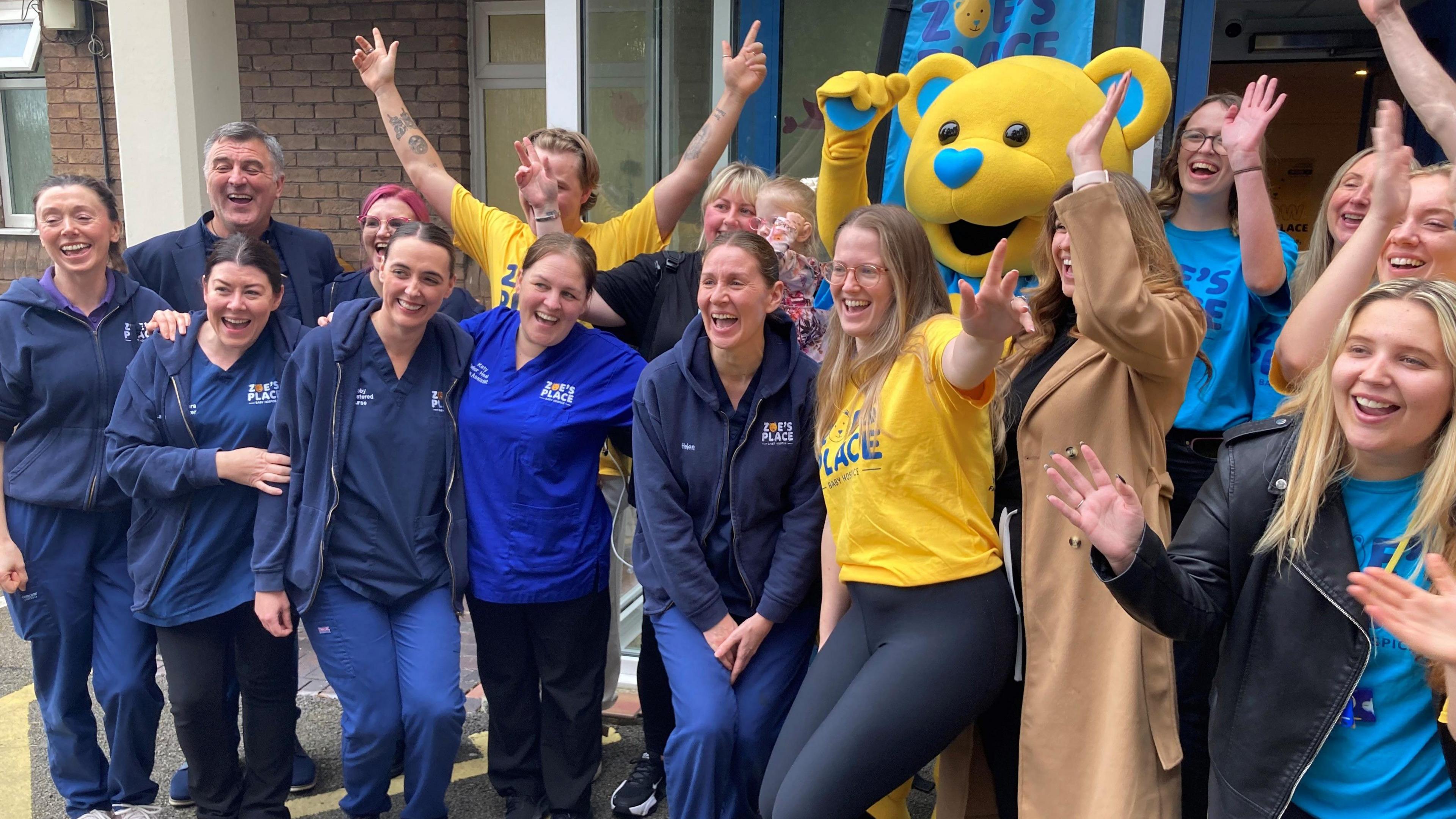 A group of nurses, some in uniform, and fundraising staff in blue and yellow Zoe's Place branded t-shirts, as well as a mascot in a yellow teddy-bear costume, smile and cheer 