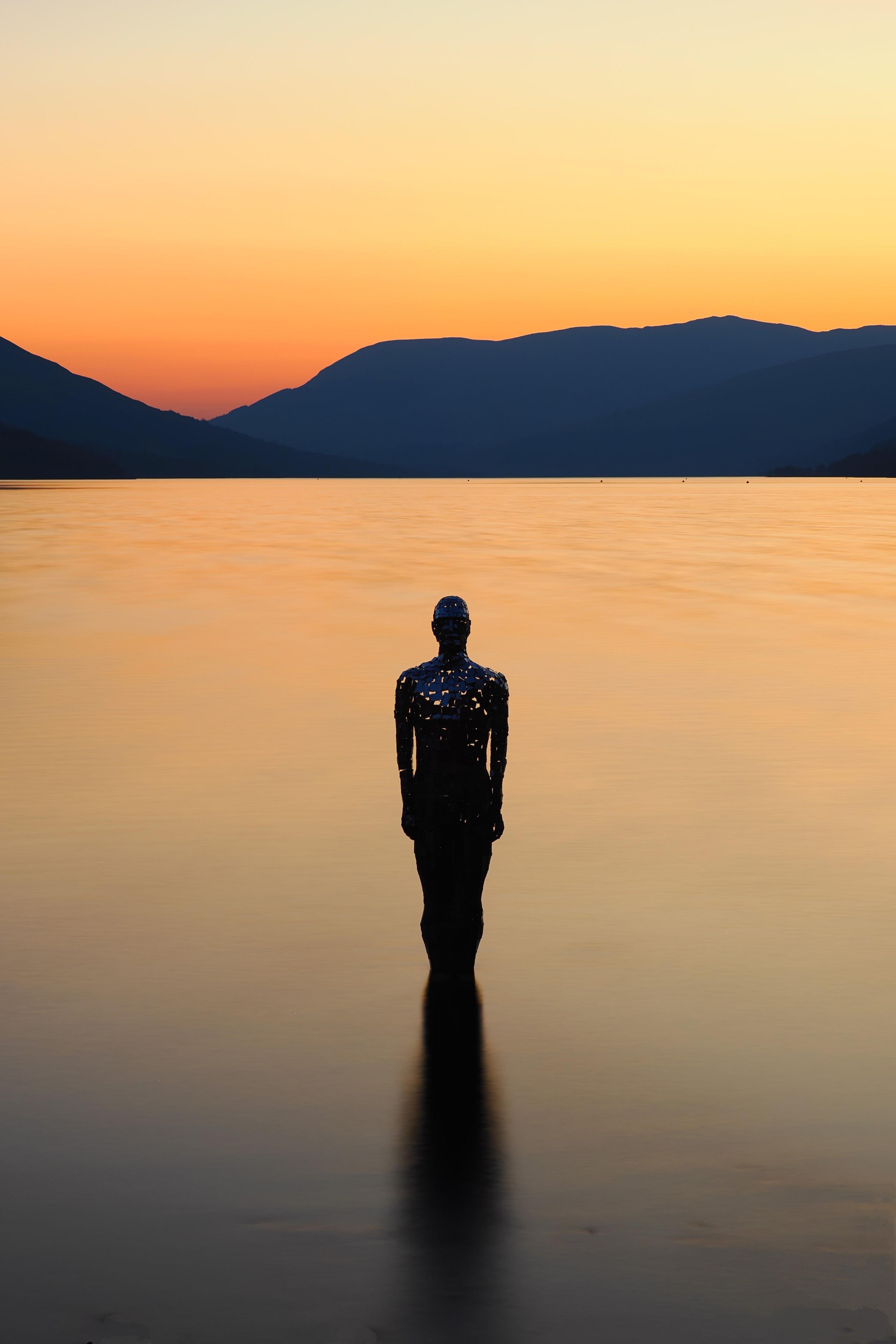 A stainless steel sculpture of a man in a loch, under an orange sky.