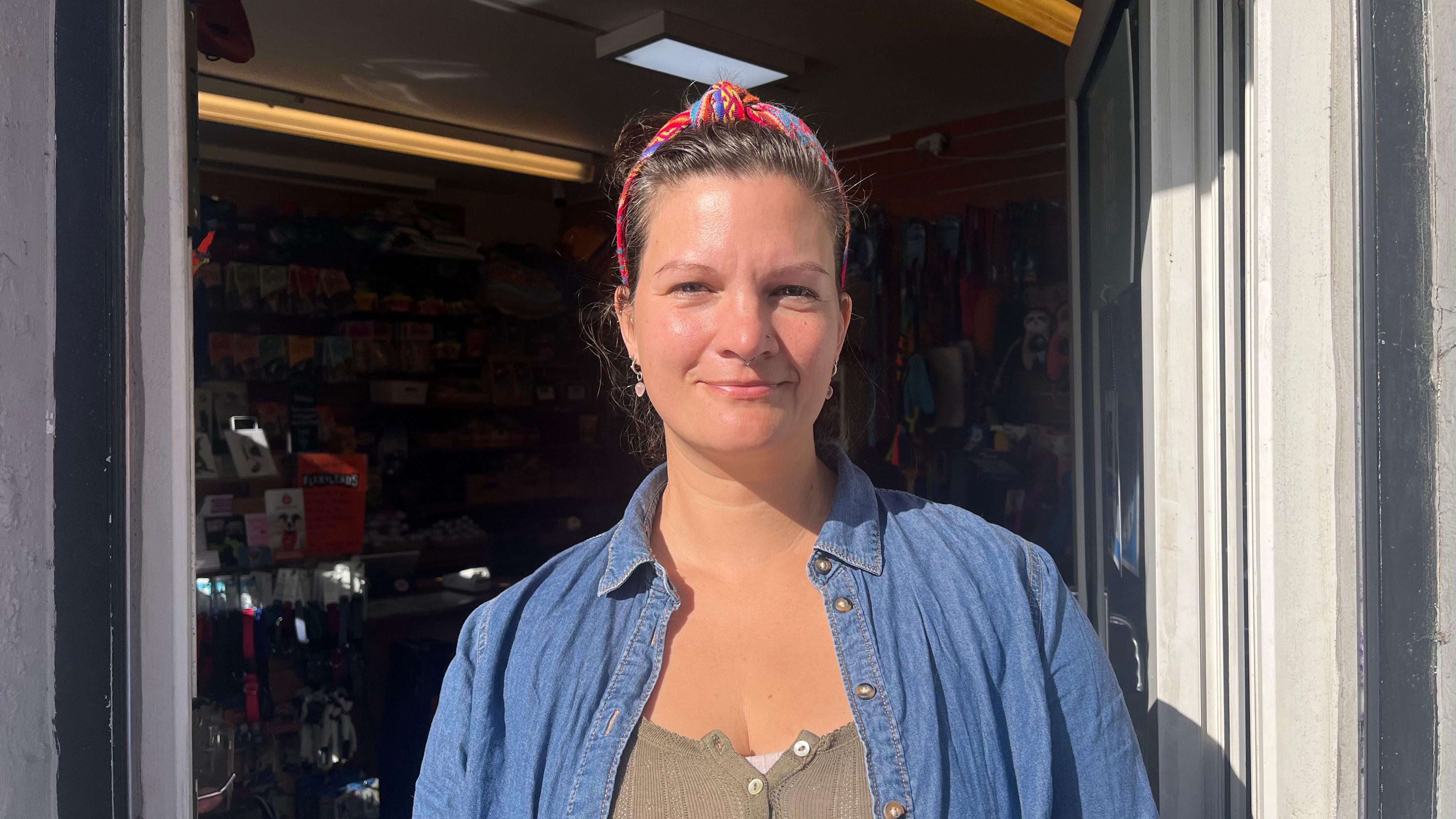A woman wears a colourful headband and a blue denim shirt over a khaki blouse, in the entrance of a shop. The sun is lightly shining on her face.