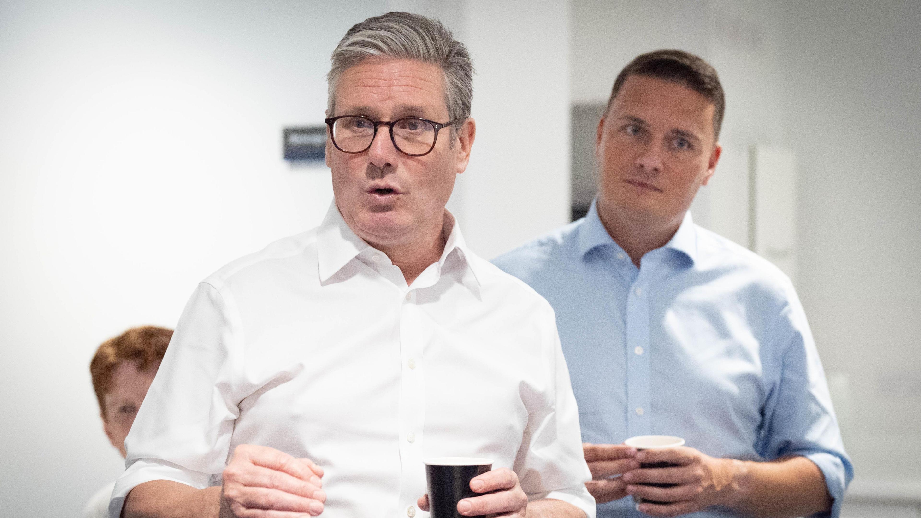 Keir Starmer, the prime minister, and health secretary Wes Streeting in a hospital. The prime minister is wearing a white shirt while Streeting wears a blue shirt. They are both holding hot drinks. 