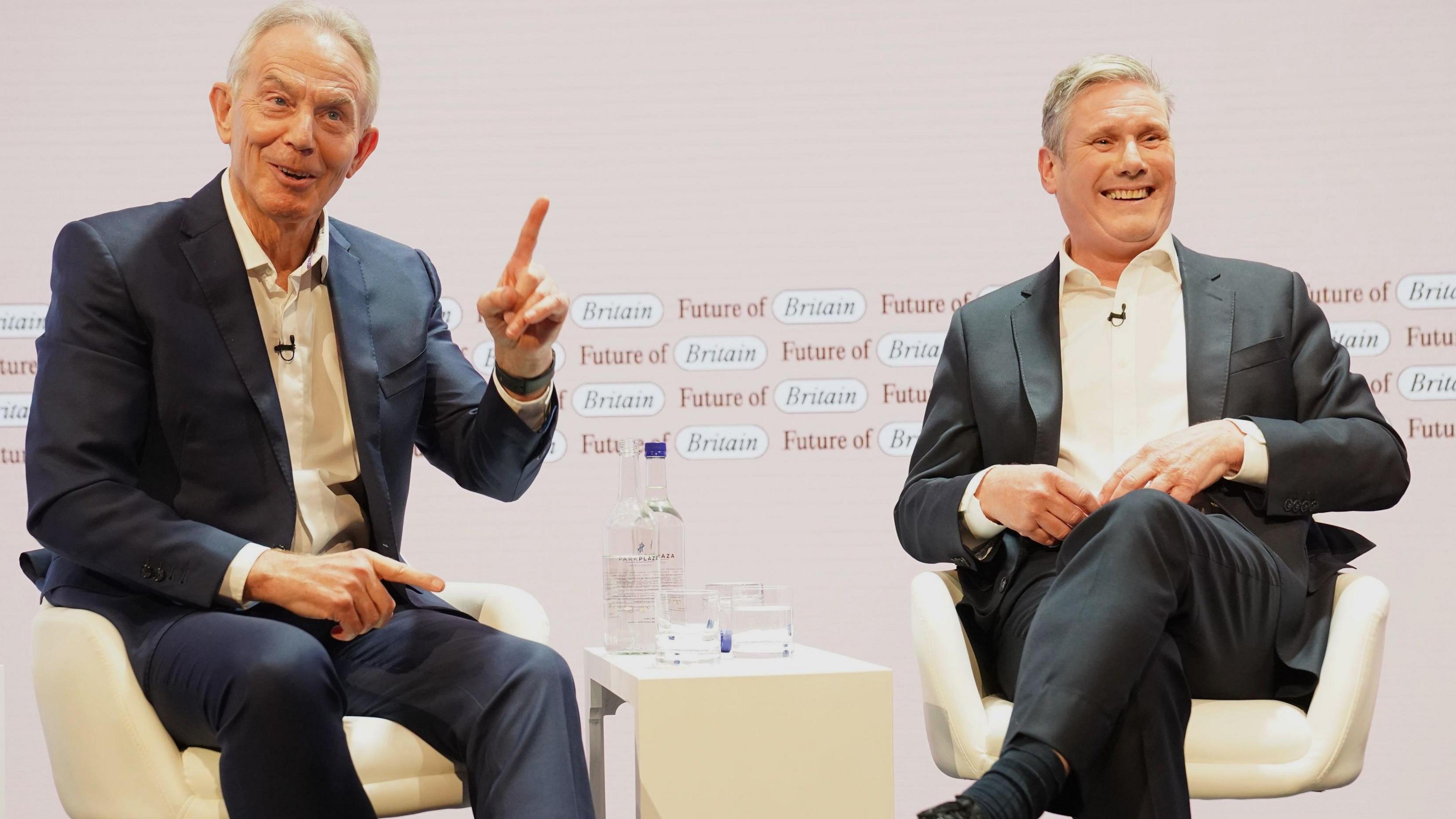 Tony Blair is sat to the left of Keir Starmer. Blair wears a navy suit and is holding his left hand in the air, pointing his finger. Keir Starmer is sat next to him. Both are laughing and appear to be in good spirits.