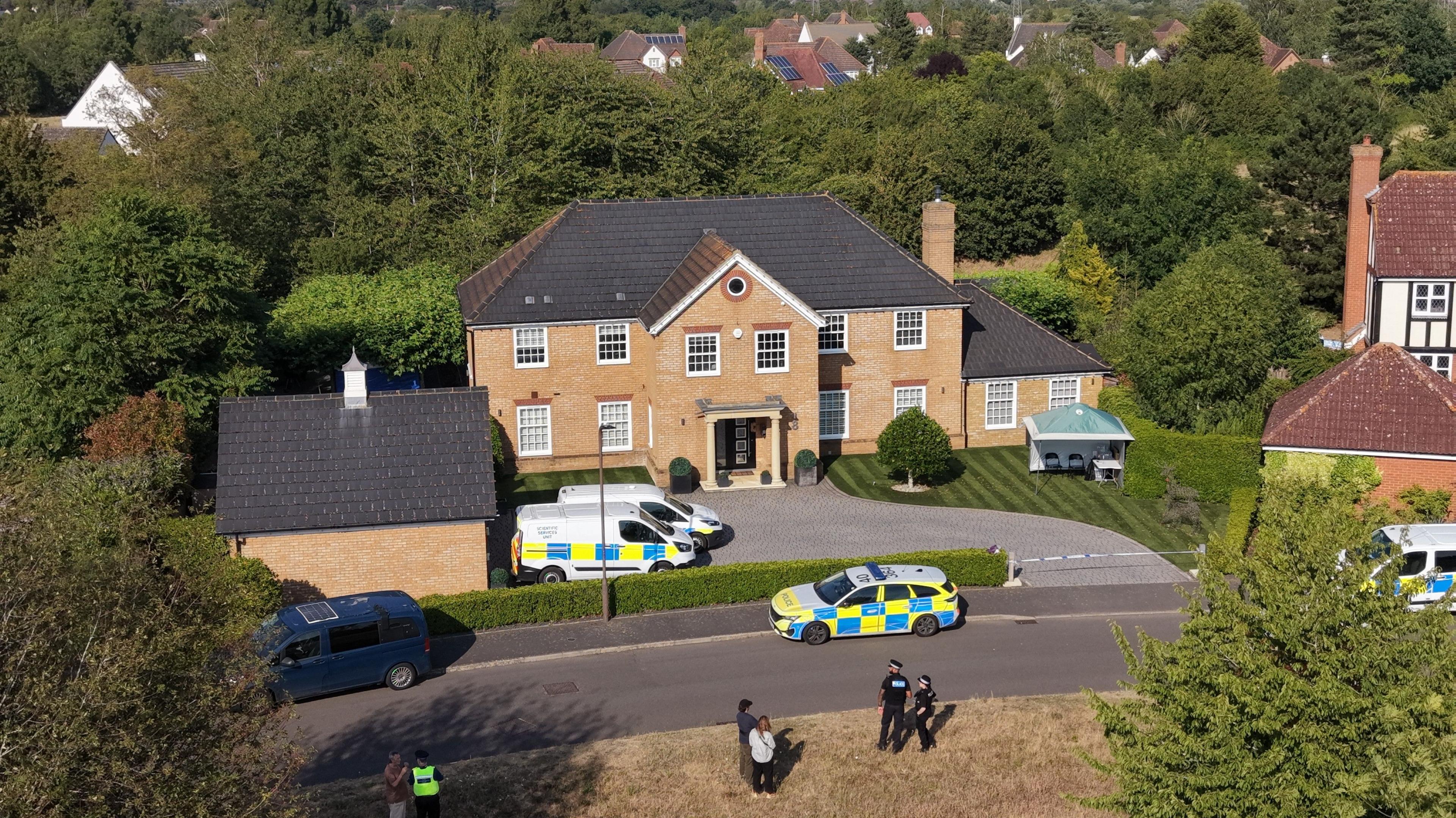 Drone view of the house in Carnoustie Drive on a bright sunny day showing police activity and vehicles. The house is a large detached property with a pillared front entrance and sweeping drive with neat lawns and sheds