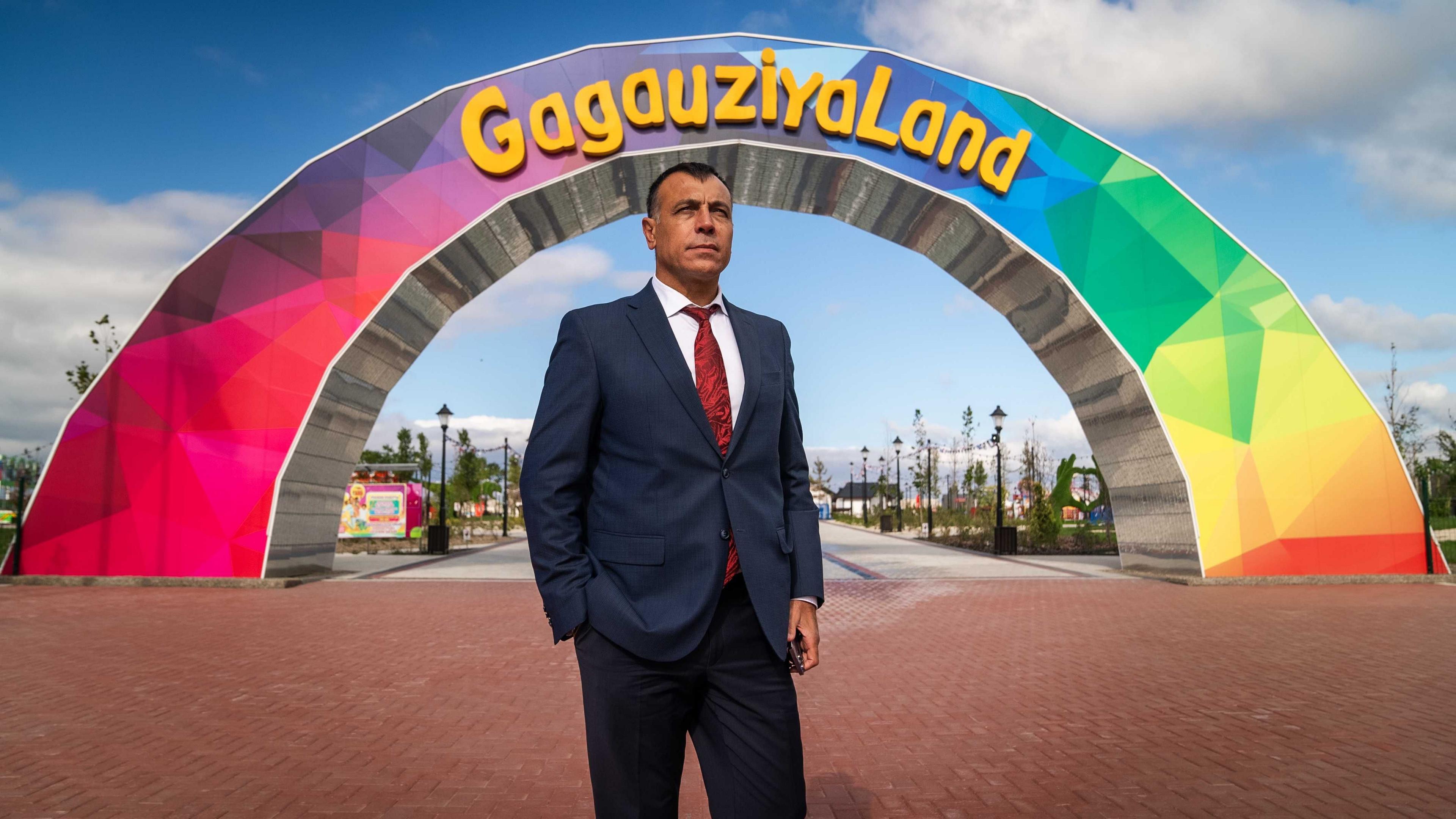 Ilya Uzun dressed in a suit, white shirt and red tie standing in front of a rainbow coloured arch that has the words GagauziyaLand written on it in yellow writing. 