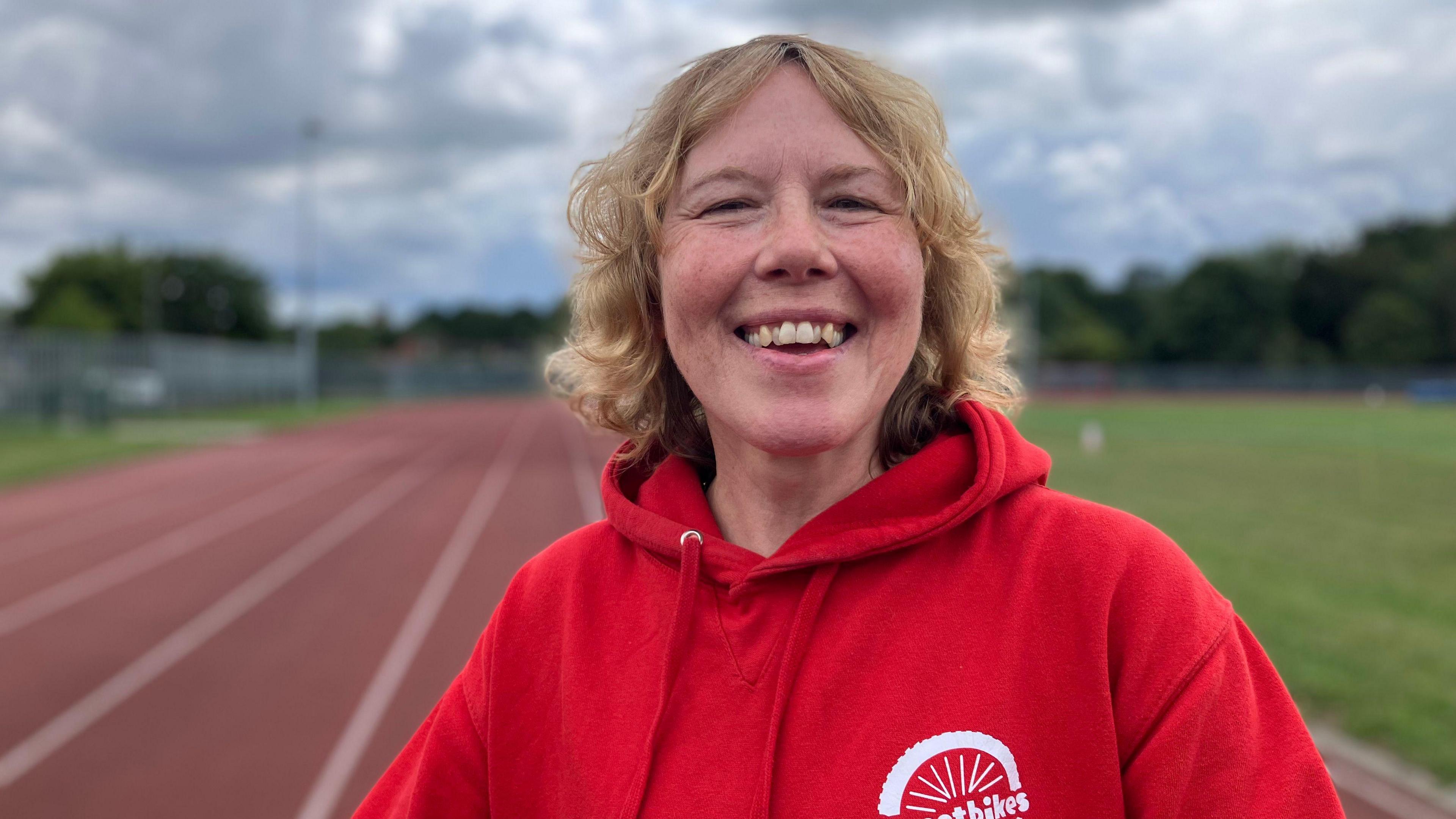 A woman with blonde hair smiles at the camera. She wears a red hooded jumper. Behind her a race track is blurred.