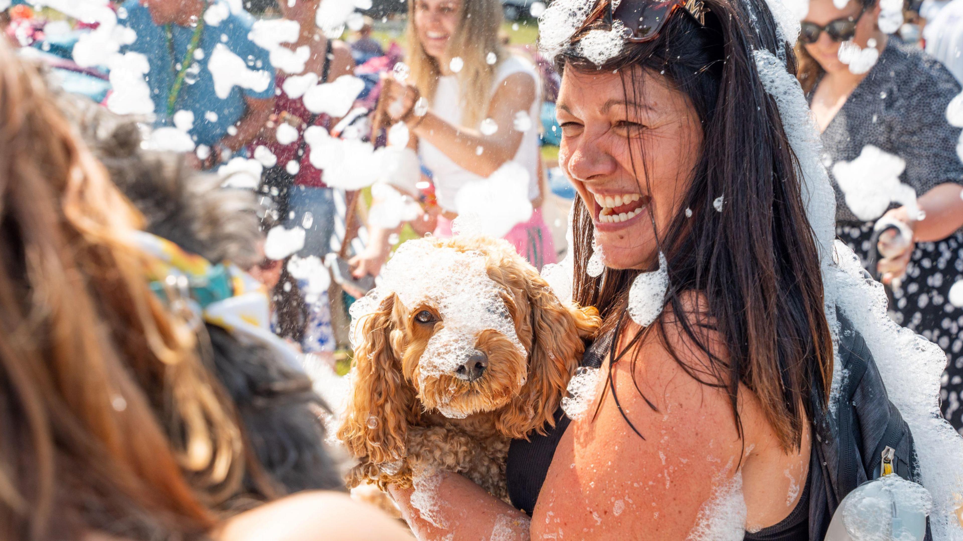 A woman is laughing while holding her dog, as they are both being covered in foam.