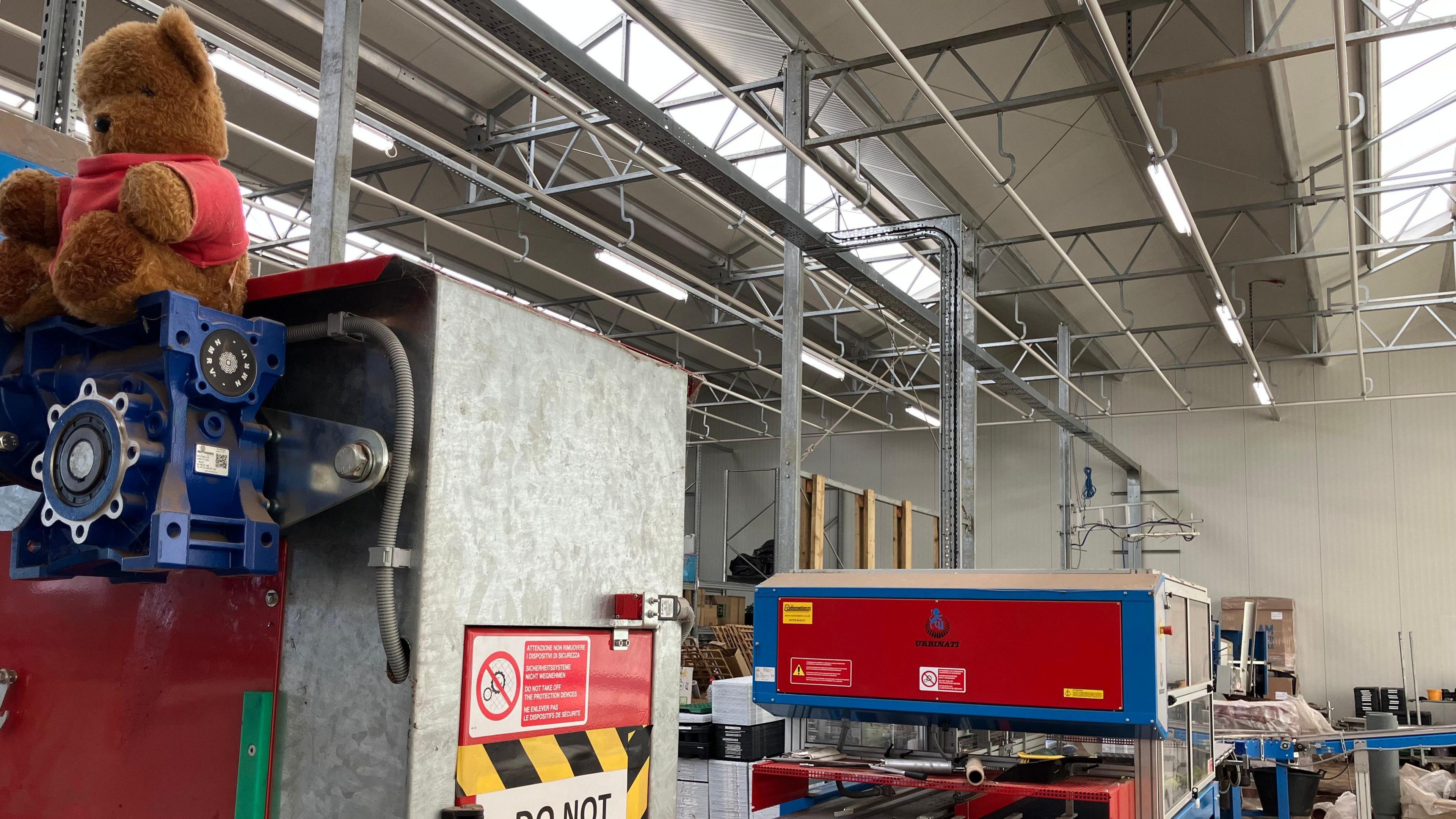 The potting machine is pictured. It includes a box with a red top and a conveyer belt. On the top of the machine is a brown bear with a red top on.