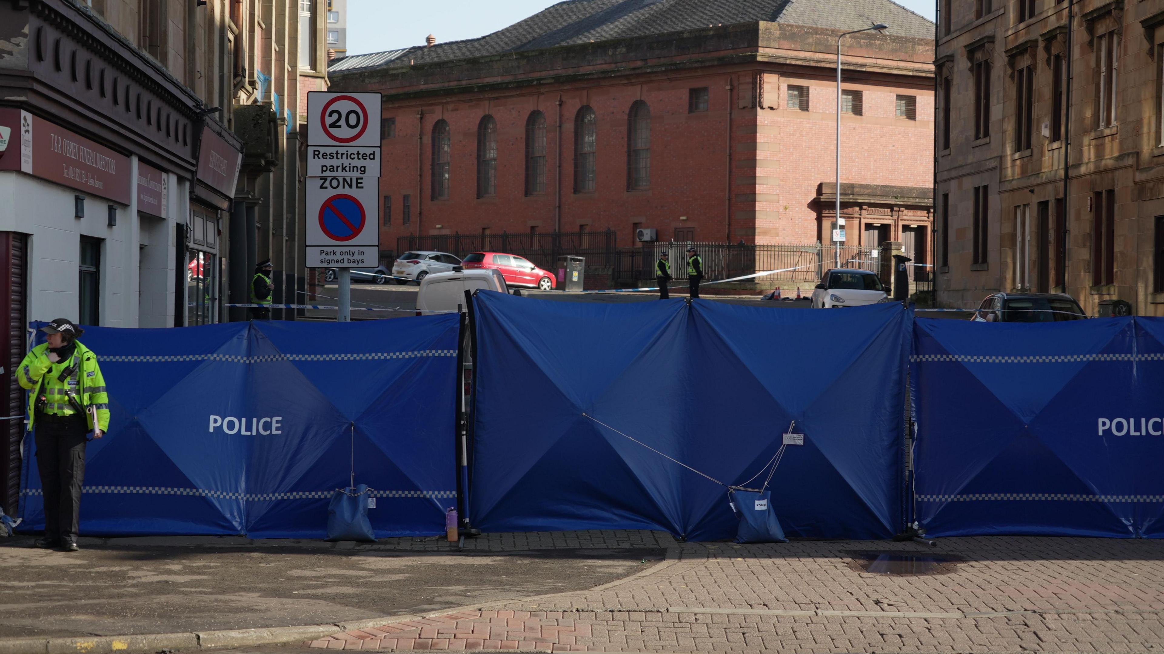 A large blue police cordon blocking the entrance to a street. Police officers are visible around it.