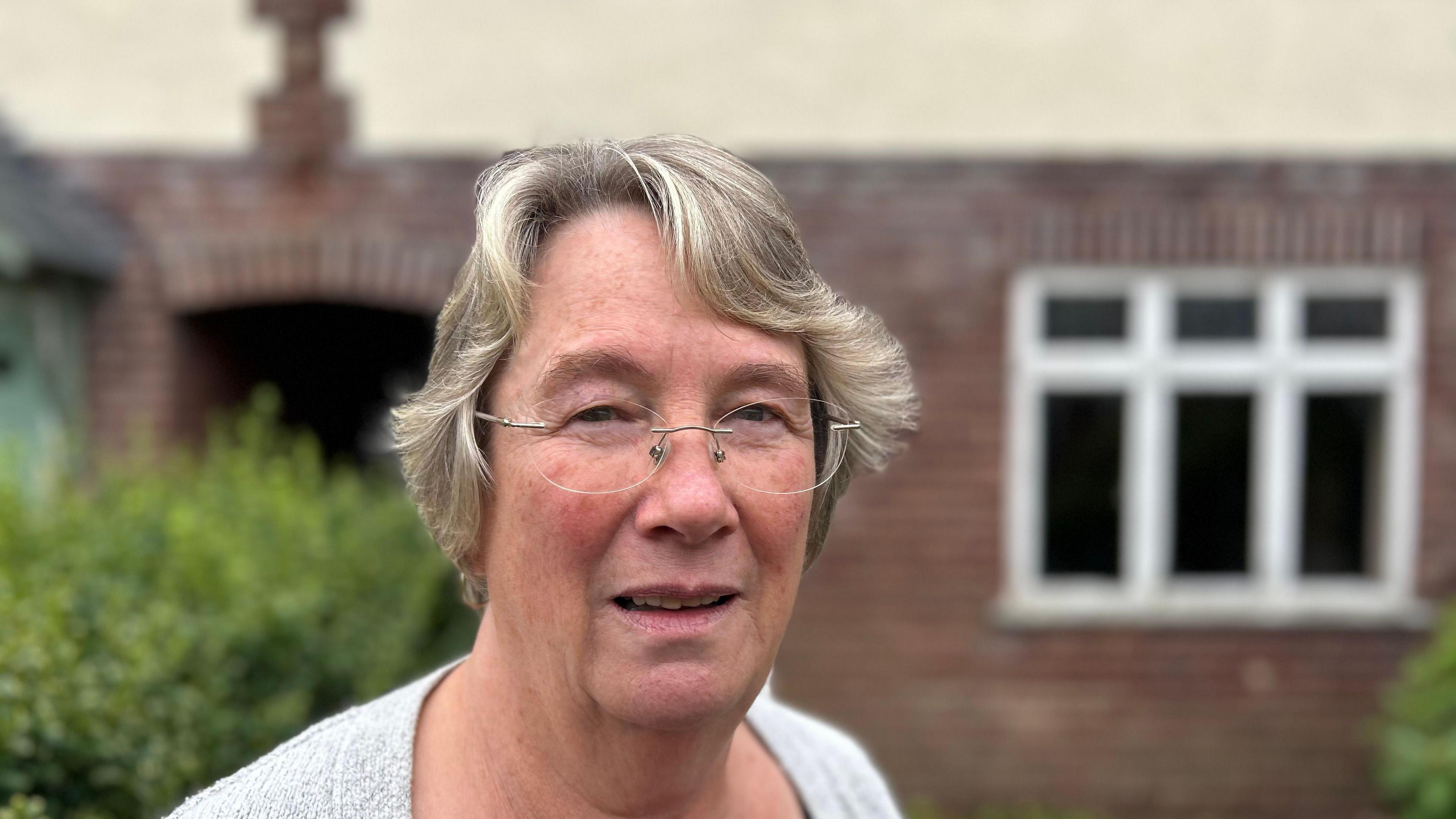 Christine Willoughby looking into the camera in front of a house. She has blonde grey hair, wears glasses and has a grey cardigan on. 