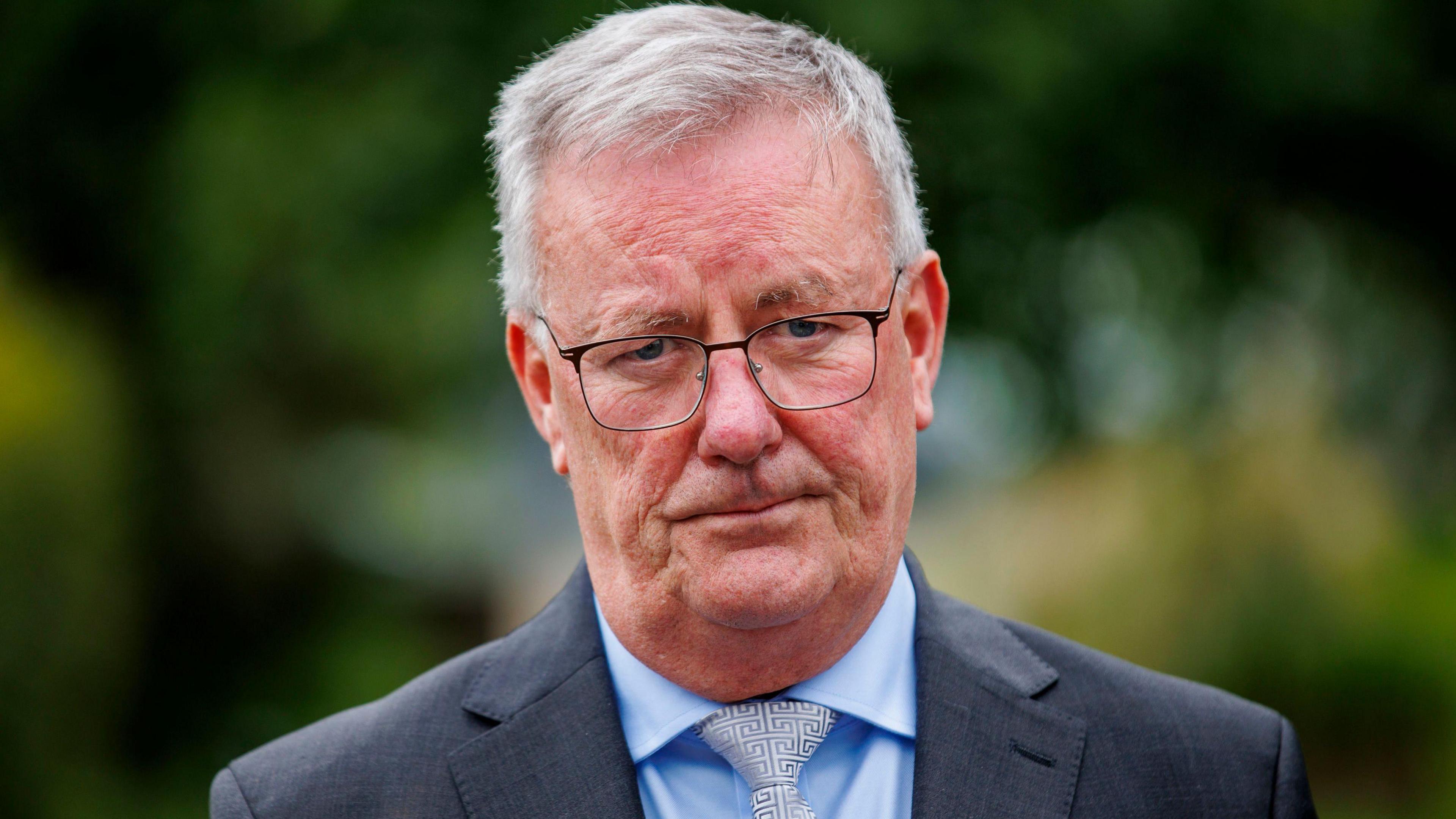 A head shot of Mike Nesbitt. He is wearing thin black glasses. He is wearing a blue shirt, tie and a grey blazer. The background is blurred.